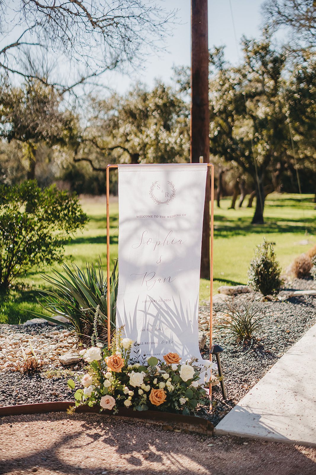 A vertical white banner on a copper stand, decorated with a floral arrangement, displays wedding text in a garden setting.