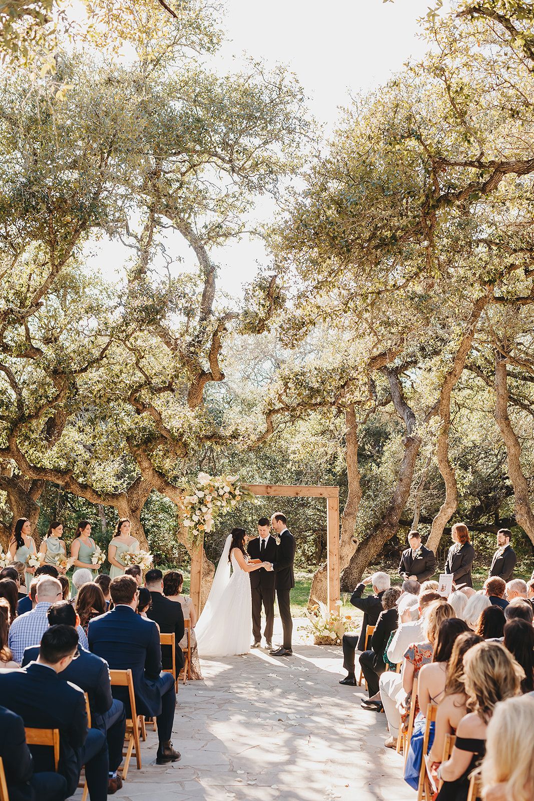 A couple stands at an altar under large oak trees during an outdoor wedding ceremony with seated guests in attendance.