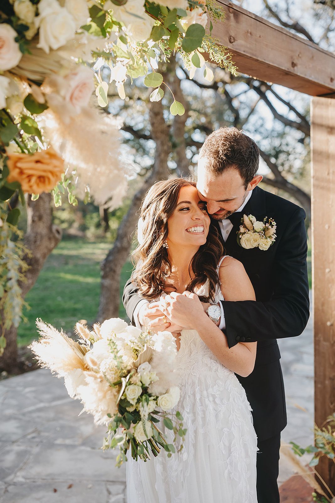 A bride holding a white floral bouquet smiles as a groom in a black suit embraces her under a decorated wedding arbor.