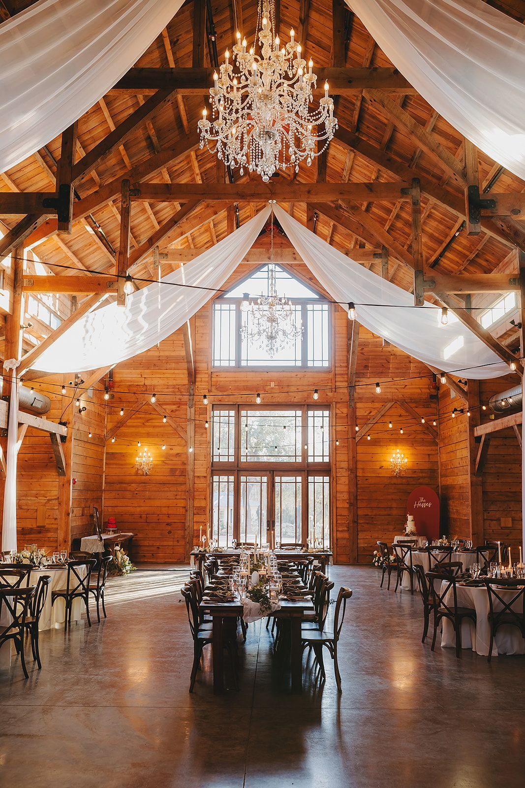A rustic wooden barn wedding venue featuring a large crystal chandelier, draped white fabric, and prepared dining tables.