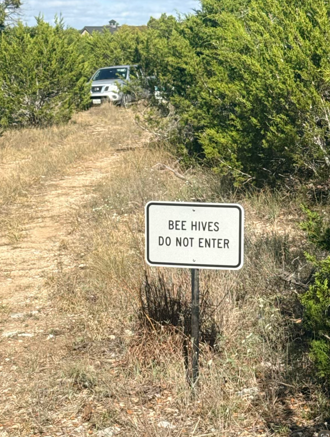 Sign reading “BEE HIVES DO NOT ENTER” beside a dirt path with a white vehicle in the background