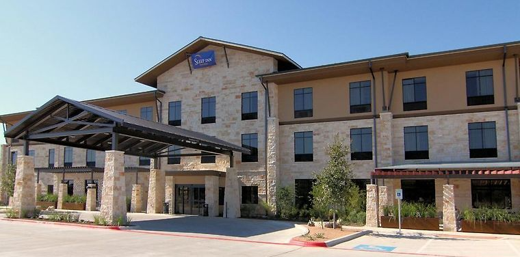 A three-story hotel with a tan stone facade, dark roof, and a covered entrance drive under a clear blue sky.