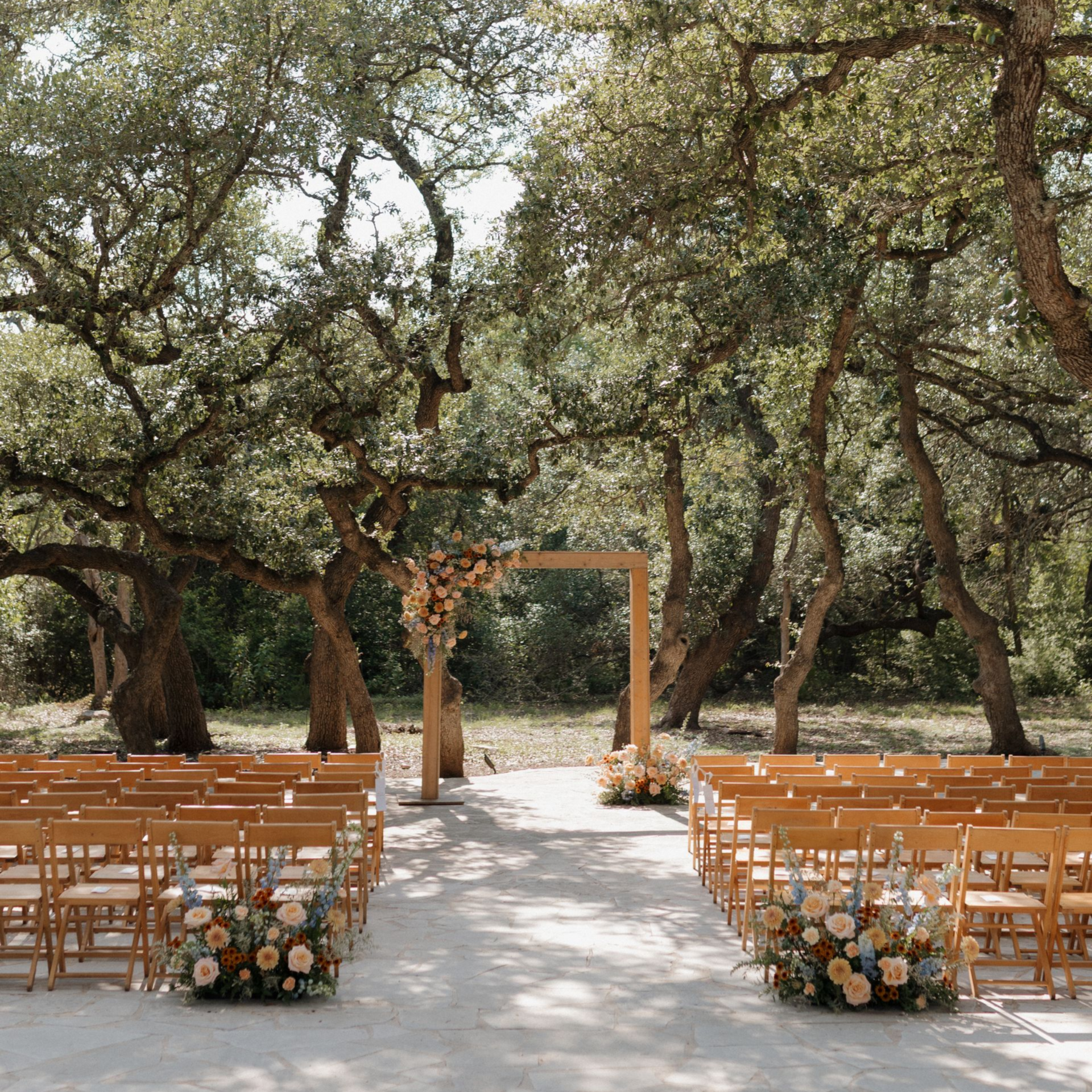 An outdoor wedding ceremony setup with rows of wooden chairs facing a wooden arbor decorated with floral arrangements.