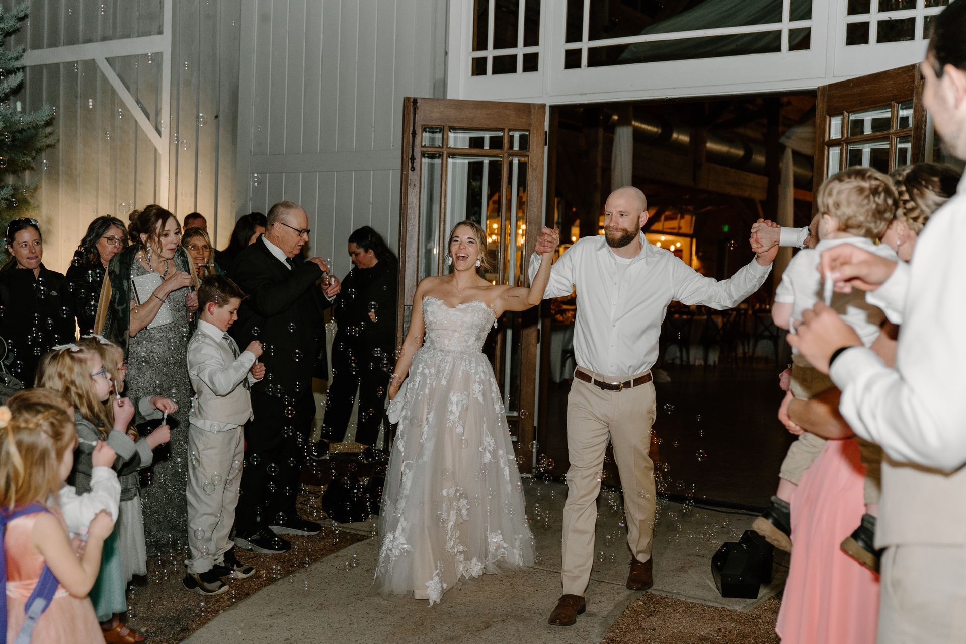 A couple exits the barn at The Addison Grove walking hand-in-hand as guests cheer amidst falling bubbles at night.