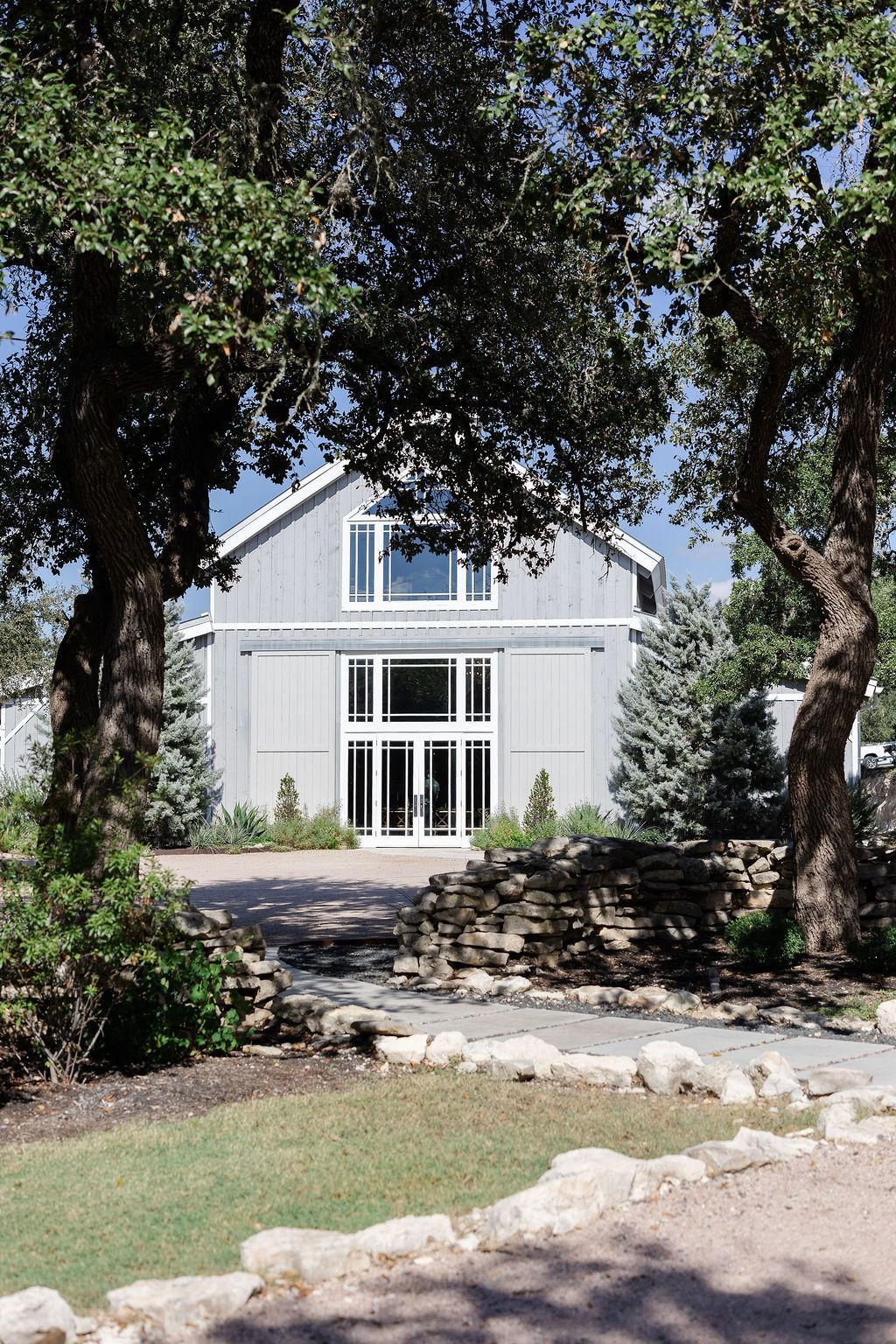 A light gray barn-style building with large glass windows, framed by two large trees and a stone pathway.