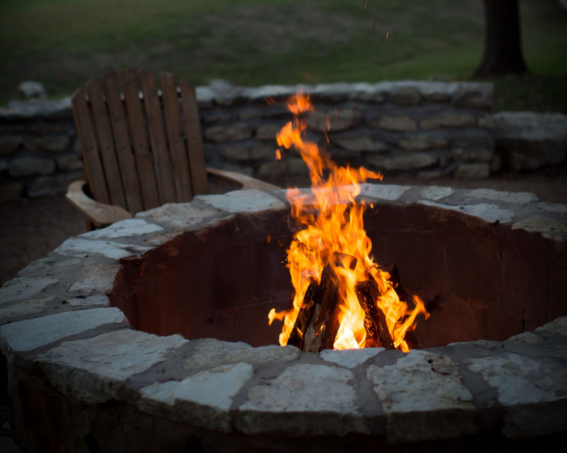 A burning campfire inside a circular stone fire pit with a wooden chair nearby in an outdoor setting at dusk.