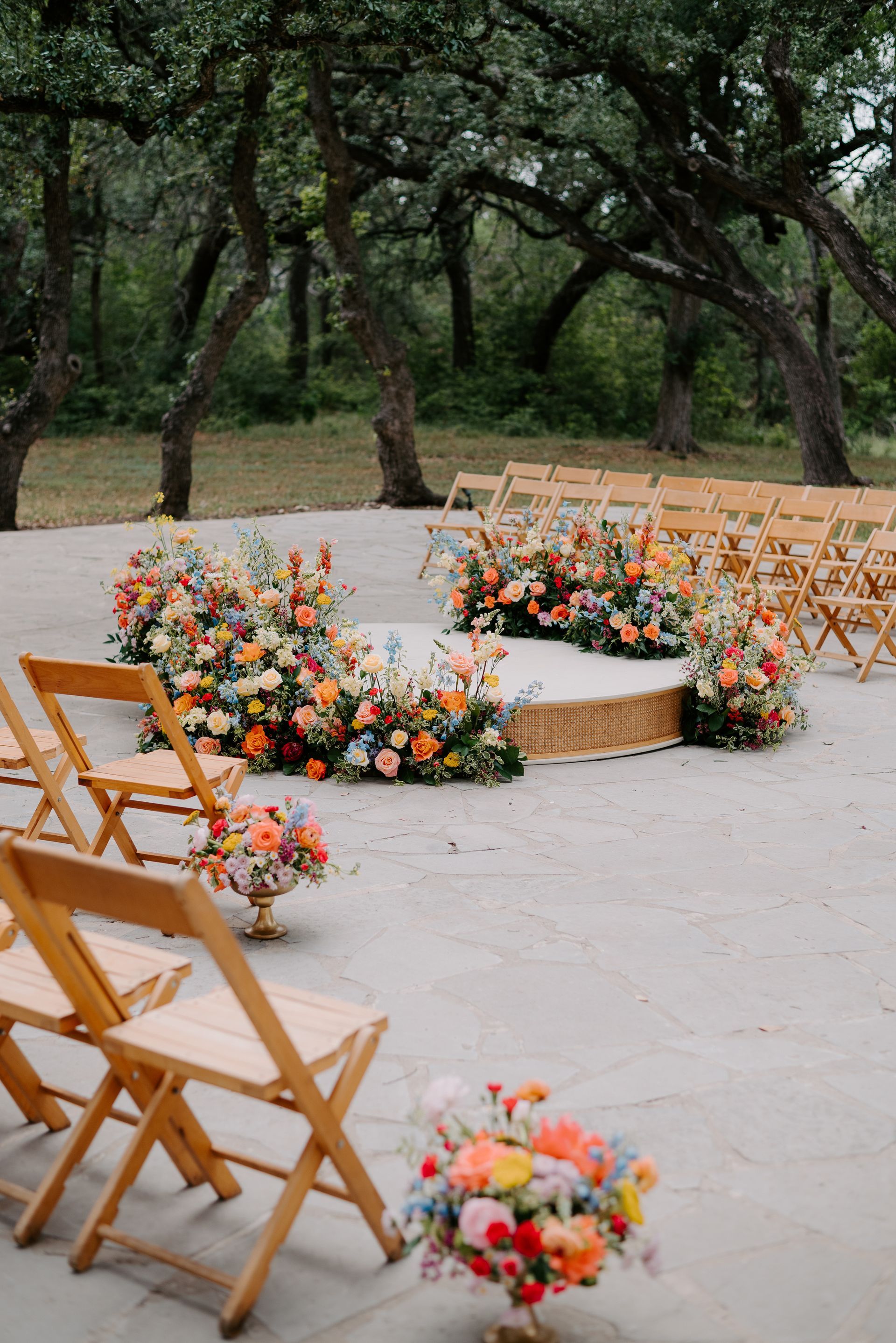 An outdoor wedding ceremony setup with rows of wooden chairs facing a floral-adorned circular platform under trees.