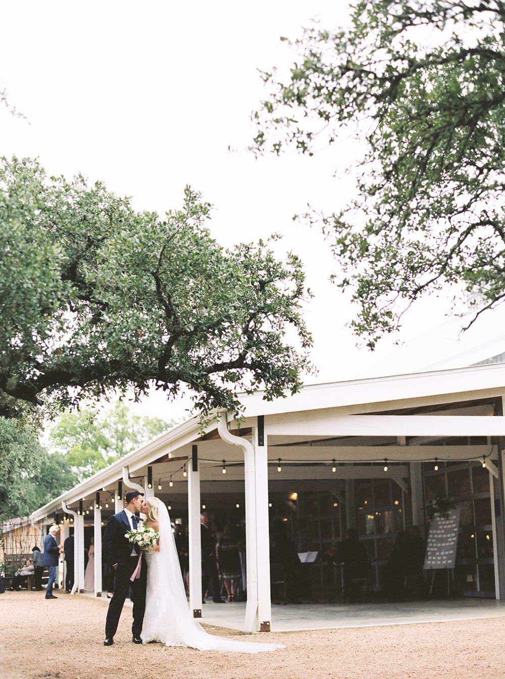 A couple in formal wedding attire kissing under a large tree next to an open-air venue building.
