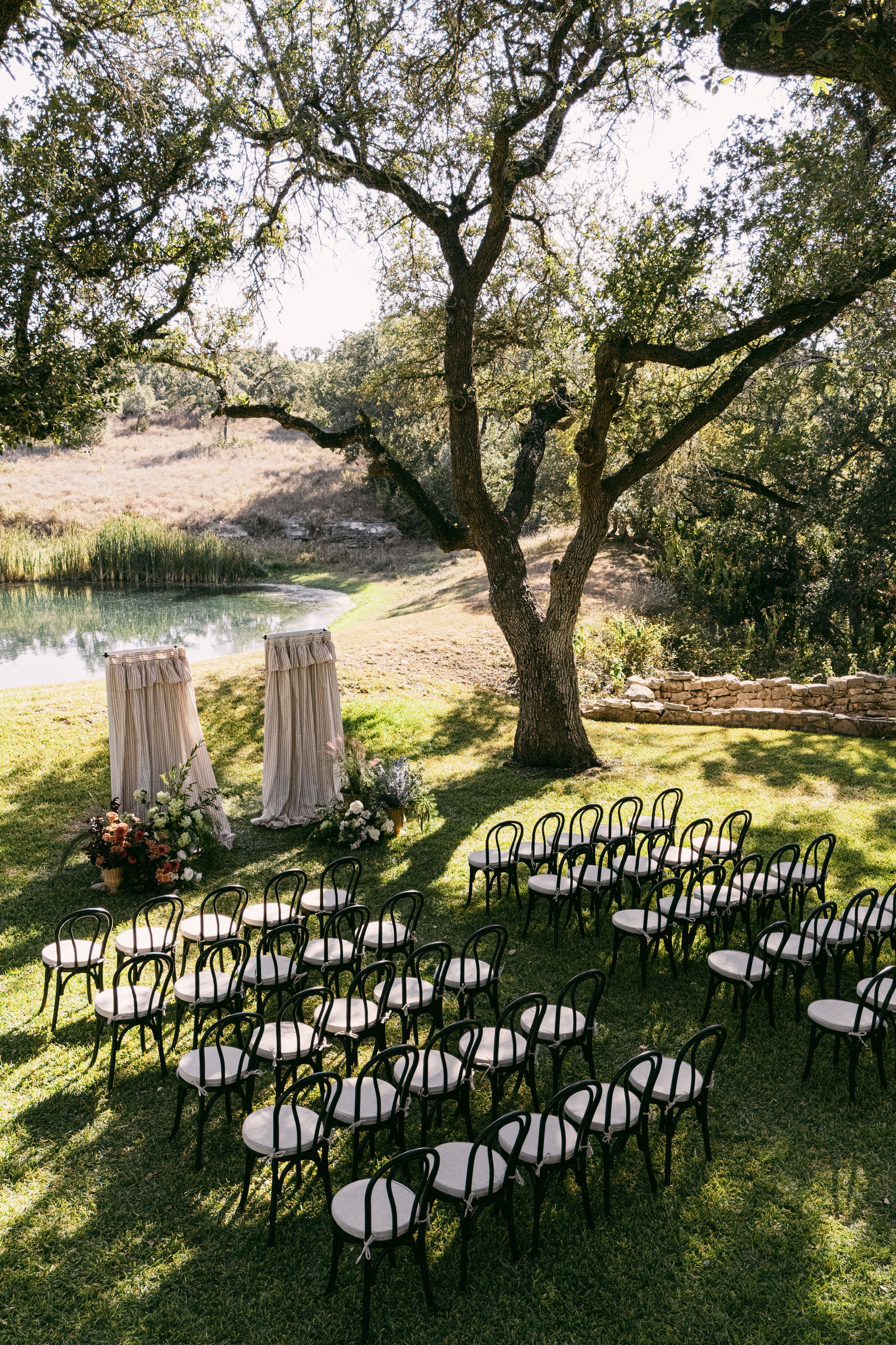 Outdoor wedding setup with white chairs on a lawn beneath trees by the water
