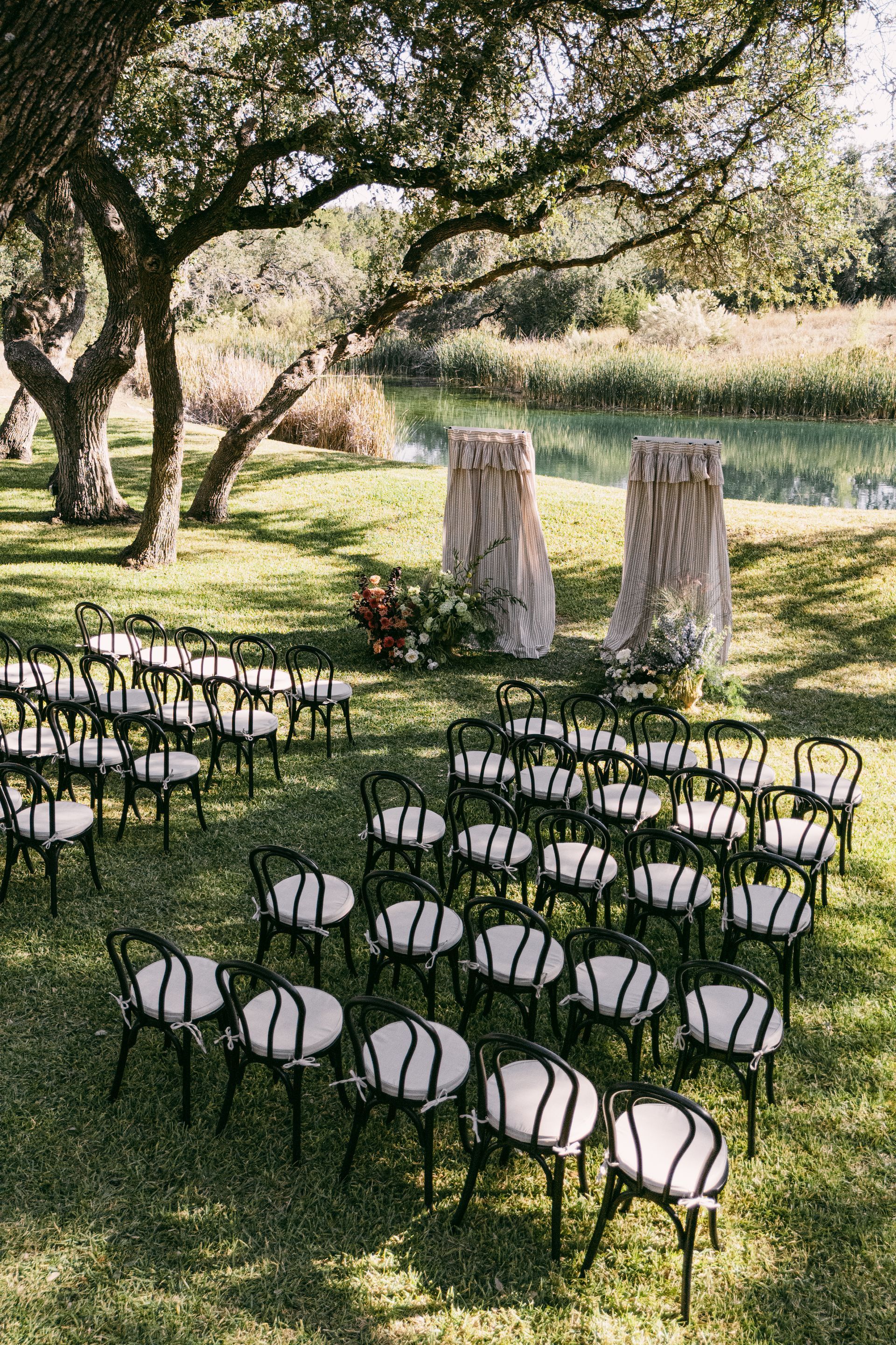 Outdoor wedding ceremony setup with rows of black chairs with white cushions on a grassy lawn near a tree and a creek.
