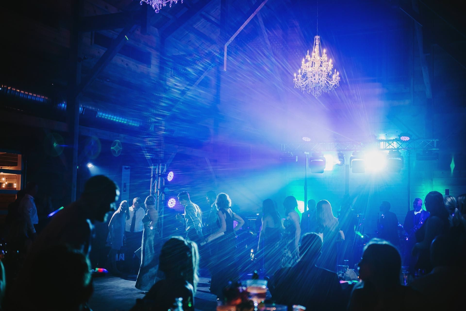 People dance in a dimly lit, blue-tinted venue with a glowing chandelier and bright stage lights overhead.