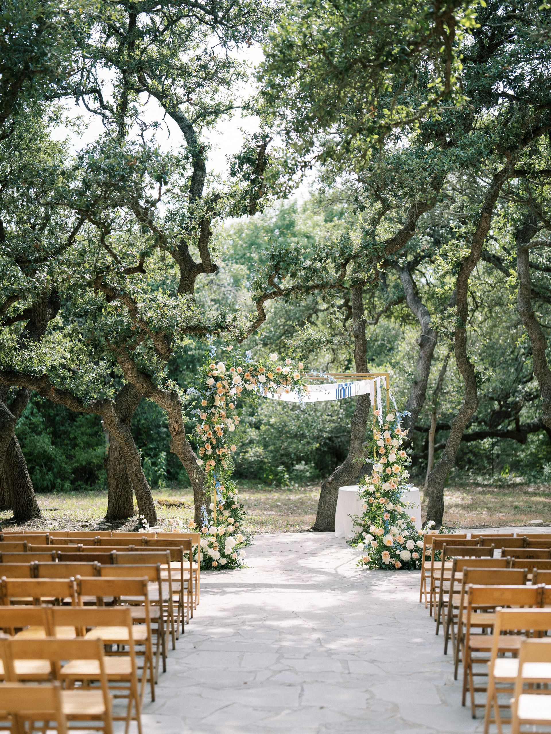 An outdoor wedding ceremony setup with wooden chairs facing a flower-adorned archway under a canopy of oak trees.