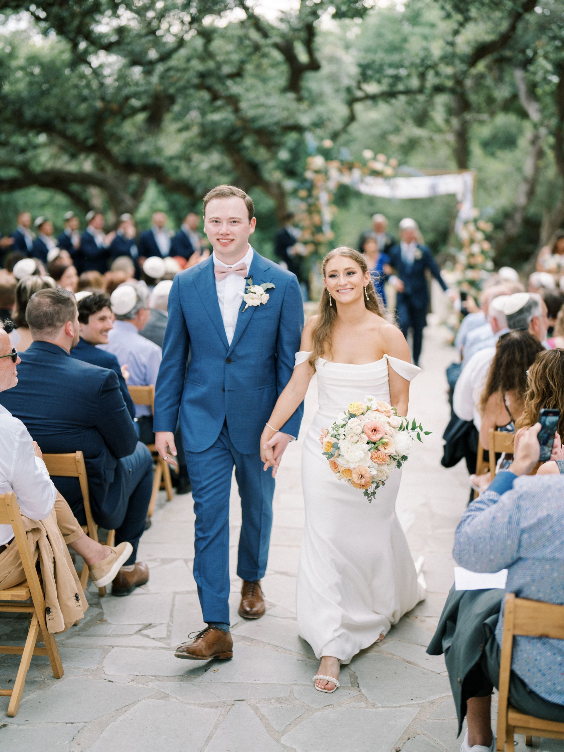 A bride and groom walk hand-in-hand down an aisle after their wedding ceremony, surrounded by seated guests outdoors.