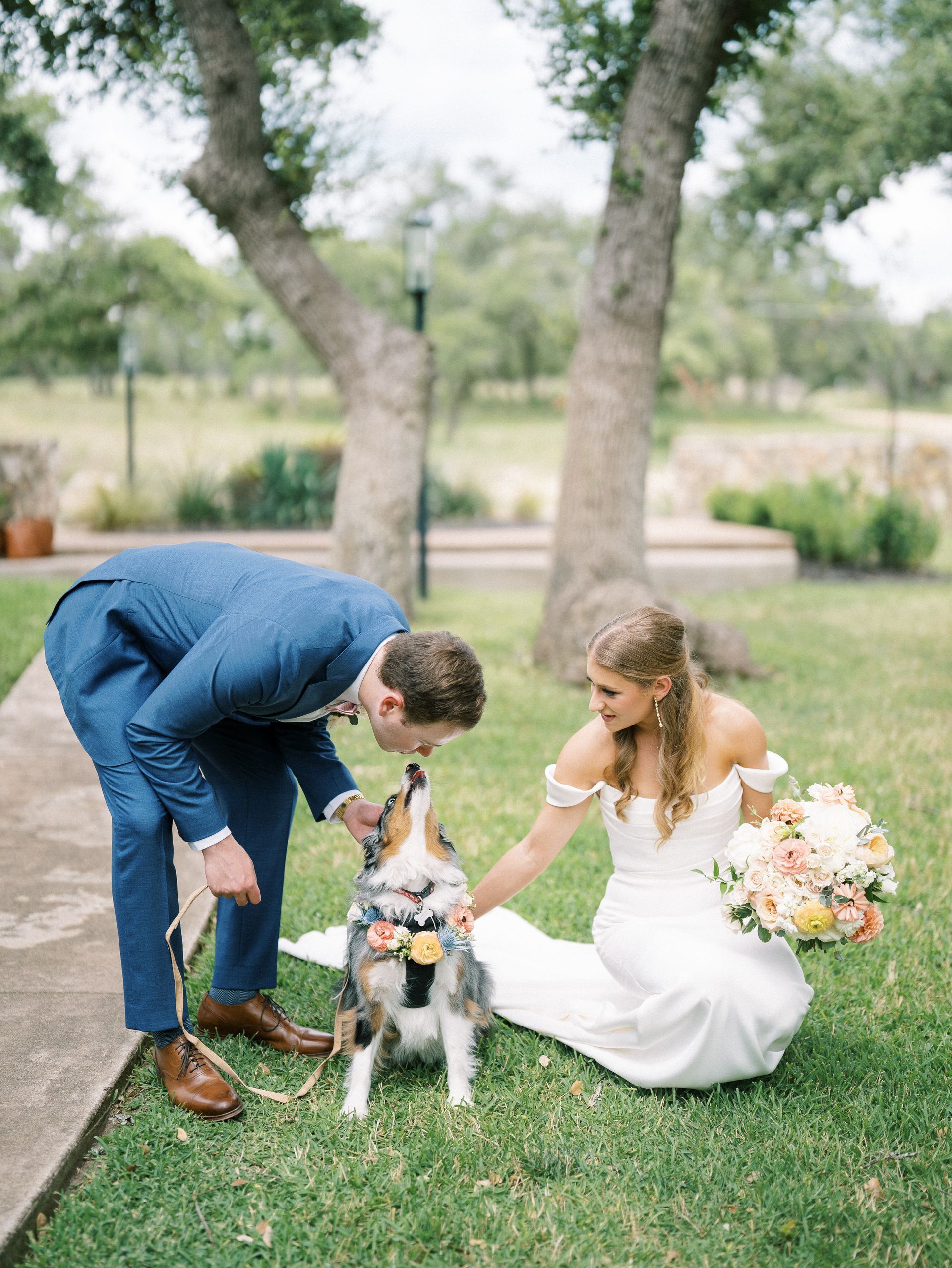 A bride and groom in wedding attire kneeling on grass, petting a dog adorned with a floral collar between them.
