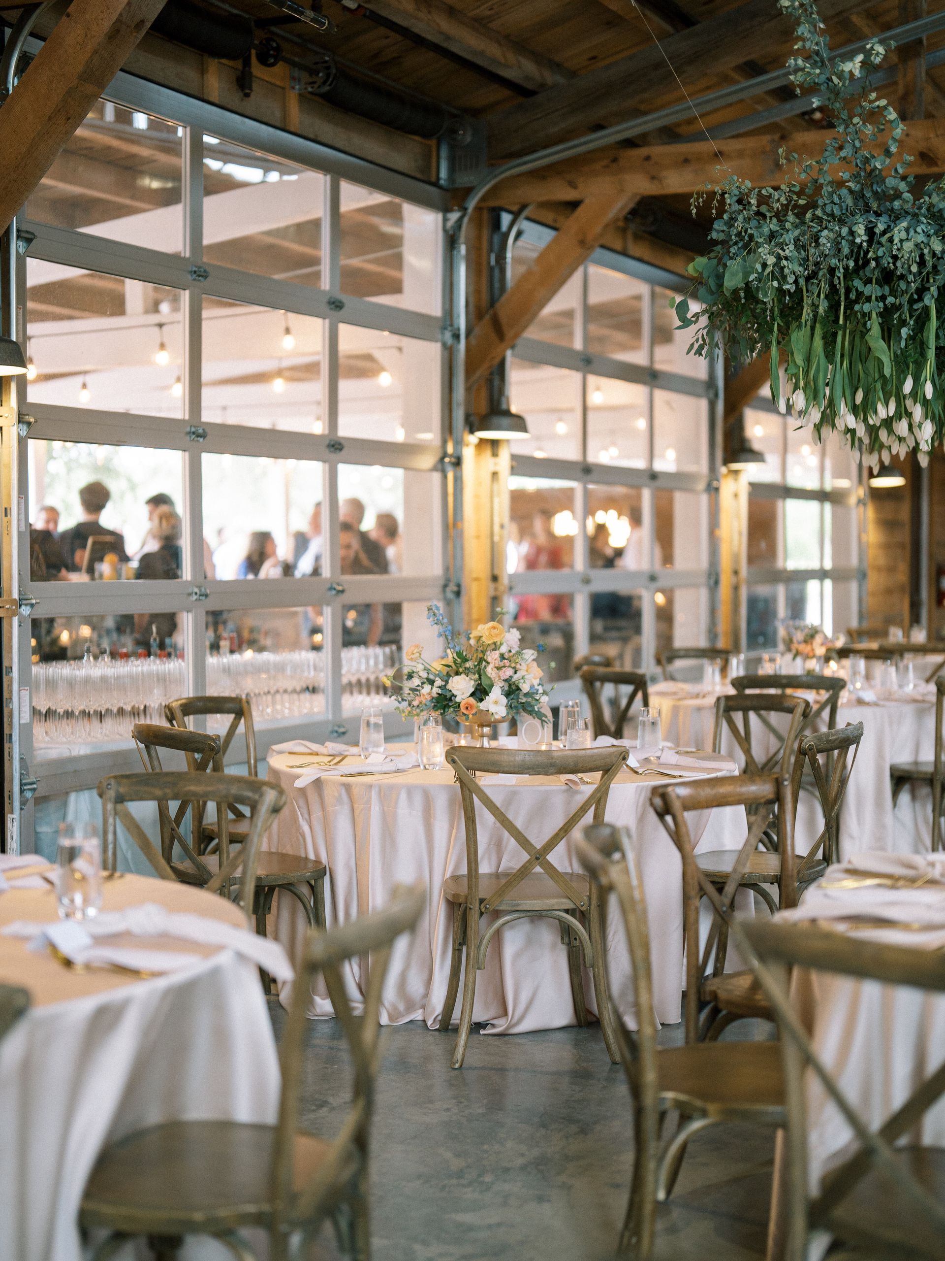 Event tables with cream tablecloths and wooden cross-back chairs inside a rustic, window-filled barn venue.