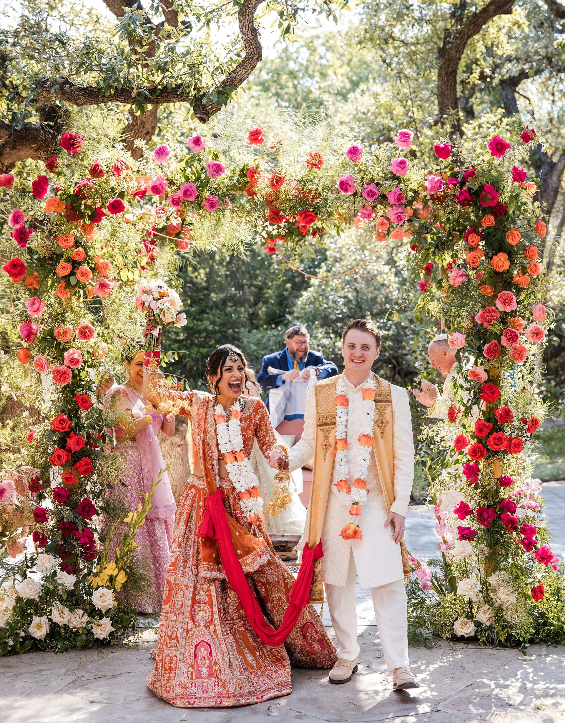 A couple stands smiling under a large floral archway decorated with vibrant pink, red, and orange flowers at an outdoor.
