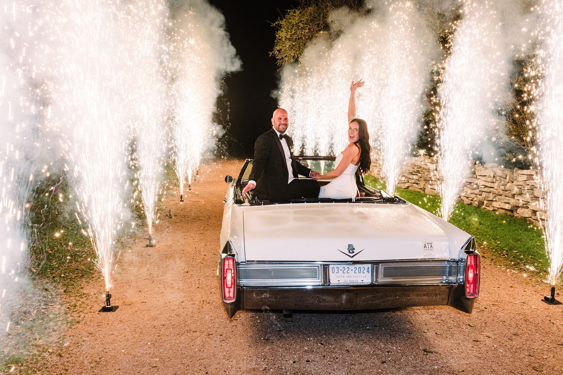 A couple sits in a white vintage convertible at night, surrounded by sparkling fireworks lining a dirt path.