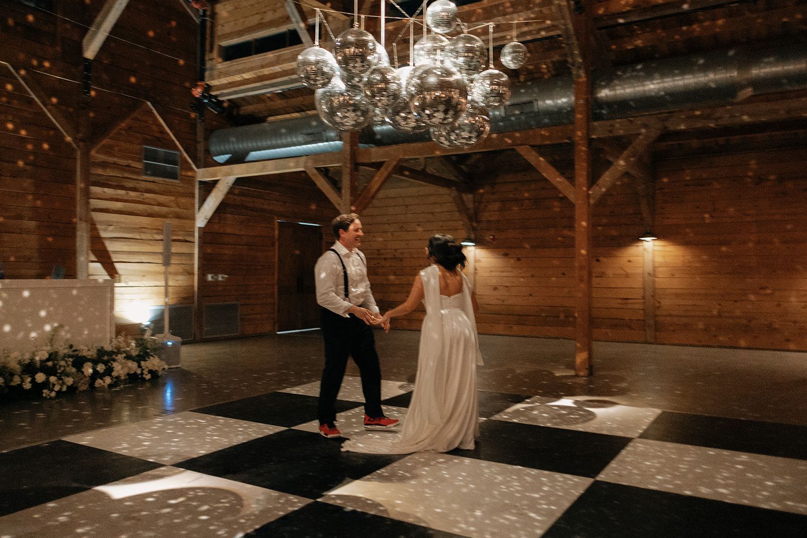 A bride and groom in wedding attire dance on a black-and-white checkered floor under disco balls in a rustic wooden barn.