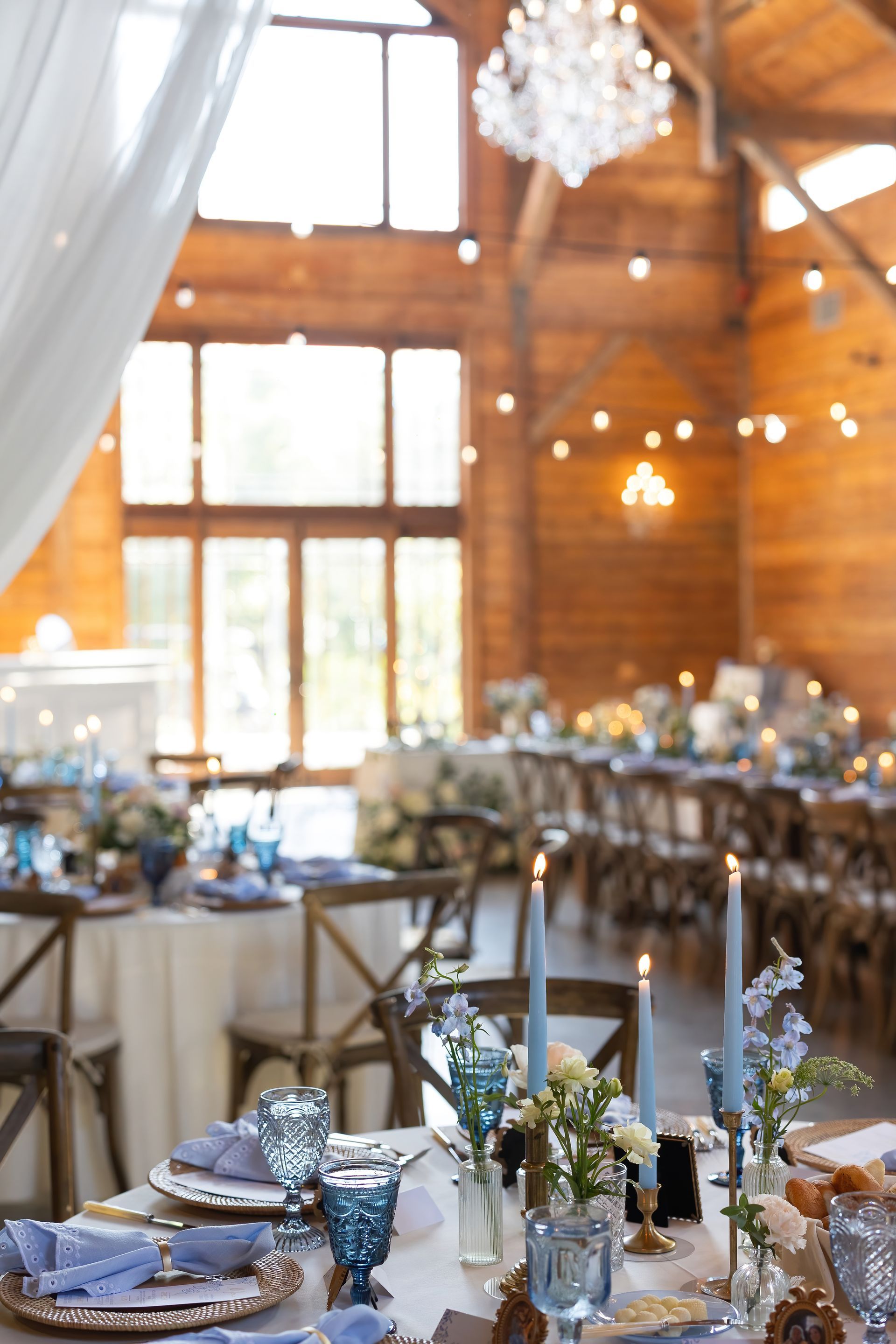 Tables set for a wedding reception in a rustic wooden barn with chandeliers and string lights.