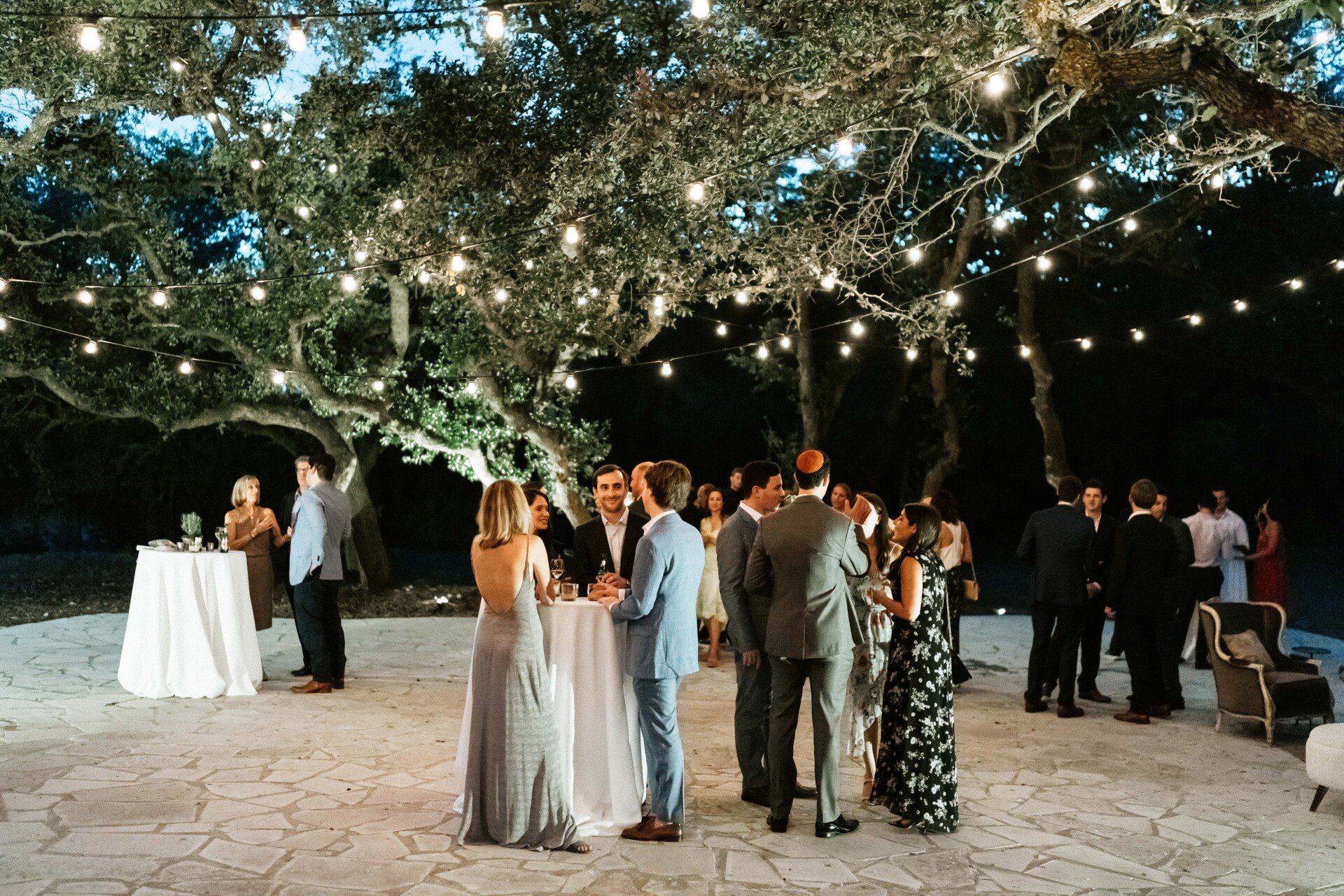 Guests socialize at a cocktail party on a paved patio beneath illuminated trees strung with warm string lights at dusk.