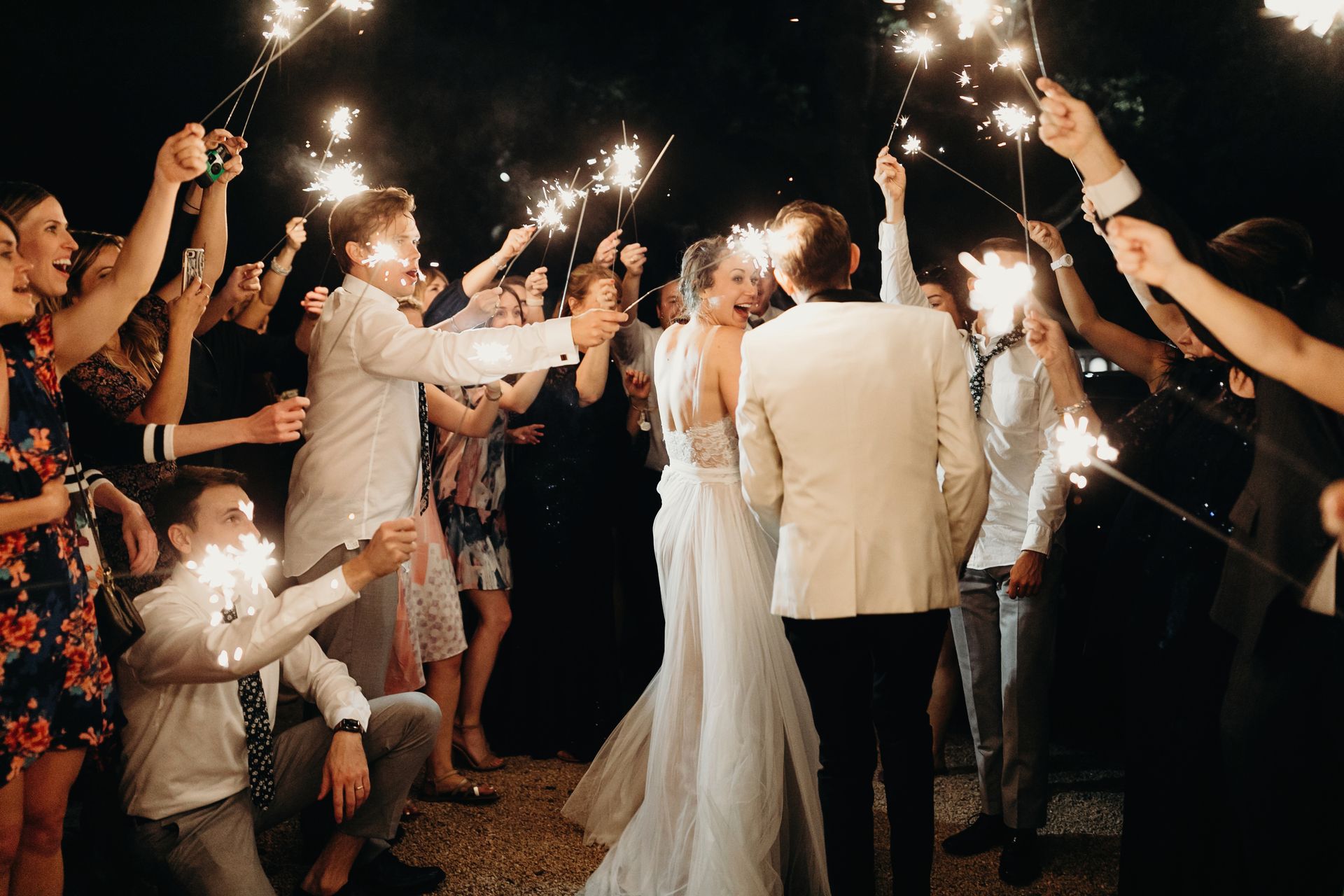 A bride and groom walk through a crowd of guests holding lit sparklers during a nighttime celebration.
