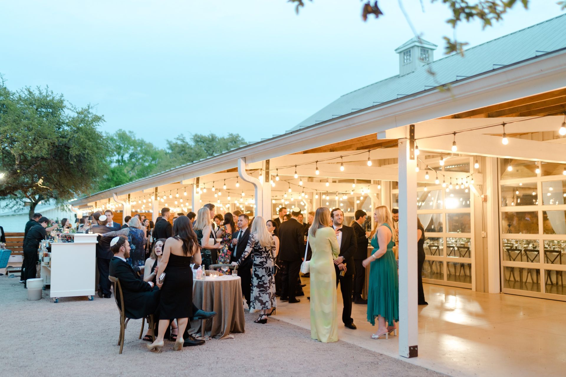 Guests mingle at a covered outdoor patio during an evening social event illuminated by string lights.