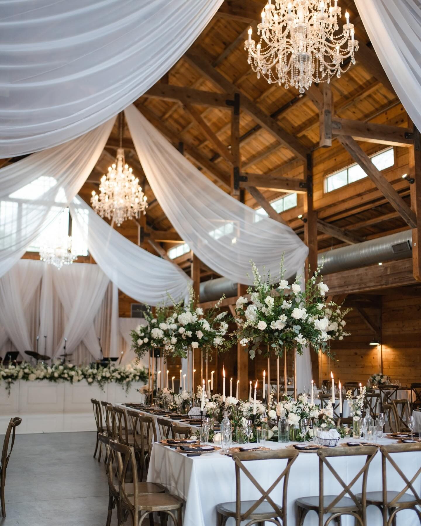 Elegant wedding reception in a rustic wooden barn at The Addison Grove with draped white fabric, crystal chandeliers, and floral centerpieces.