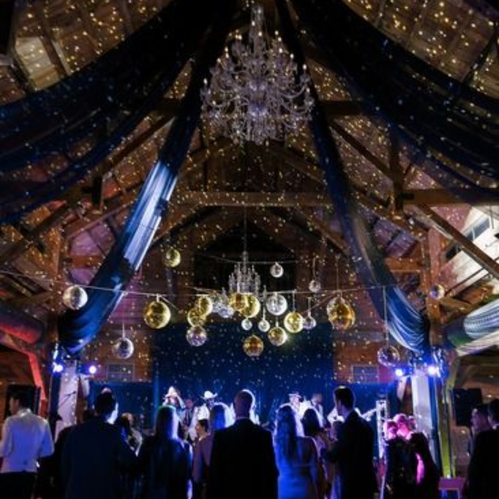 A dark, festive ballroom with high wooden beams, fairy lights, and hanging disco balls above a live band performing on stage.
