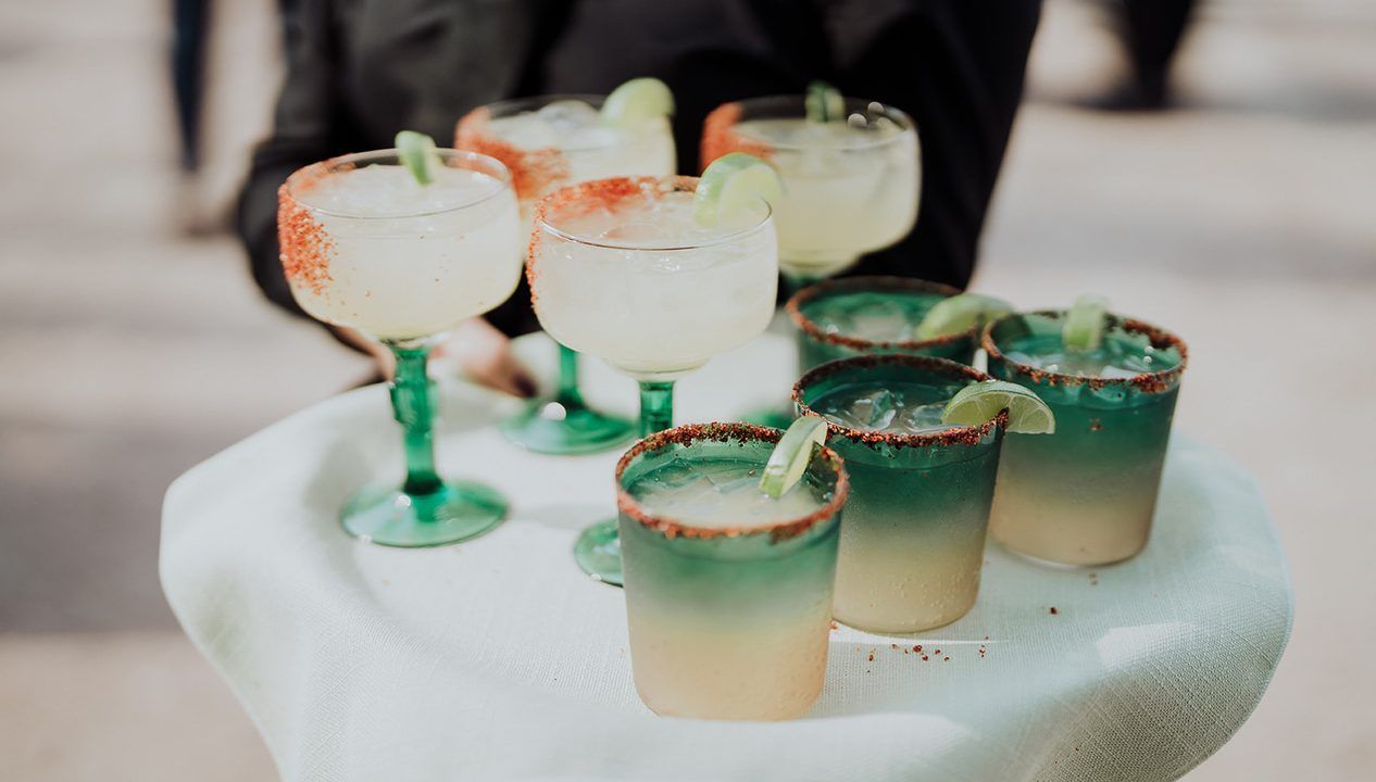 A server carries a tray with several margaritas in stemmed glasses and tumblers with salted rims and lime garnishes.