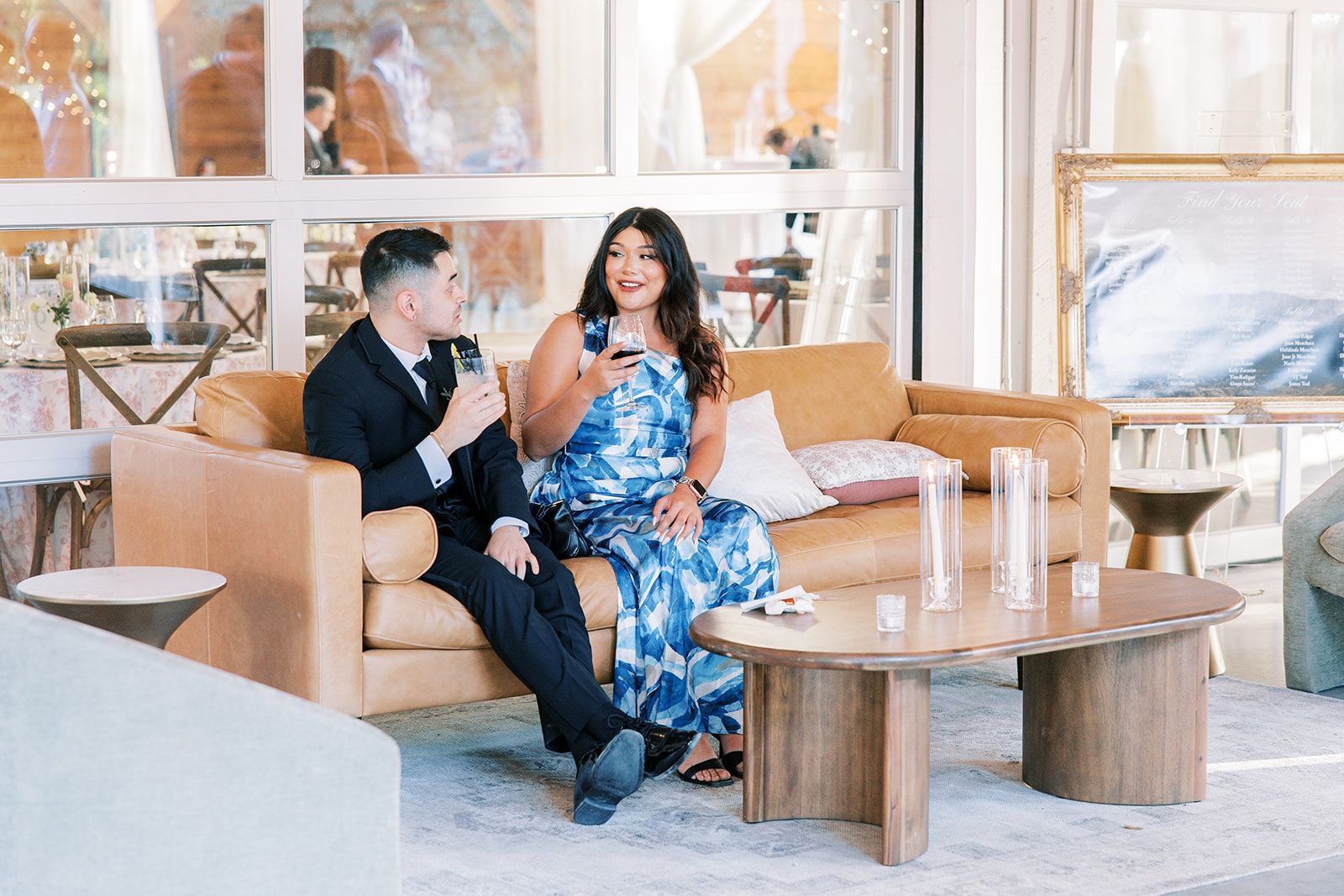 Two people sit on a tan leather sofa in a bright, modern room, smiling and holding glasses during a social gathering.