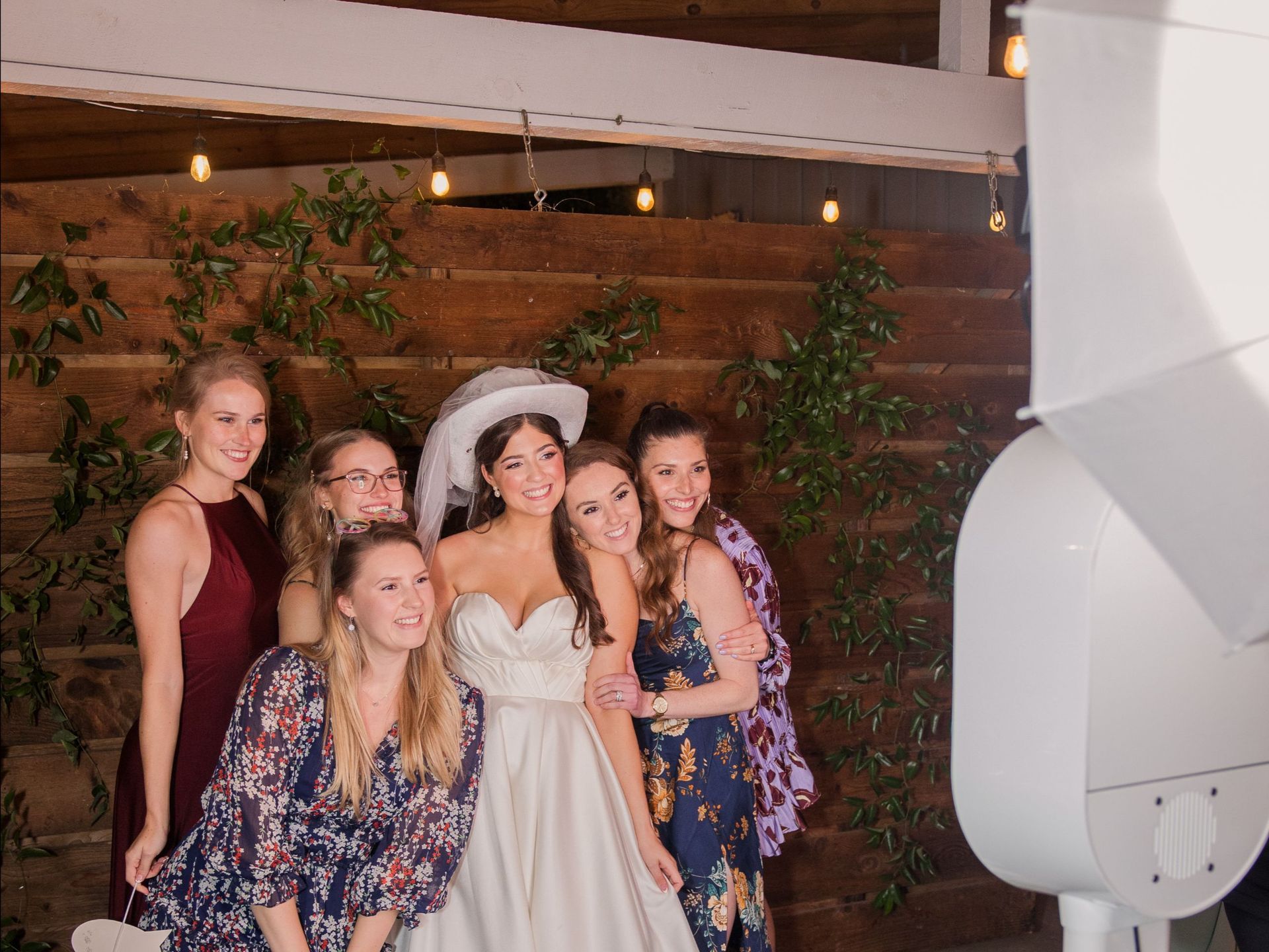 Six people pose smiling together at a photo booth against a wood-paneled wall decorated with greenery and string lights.