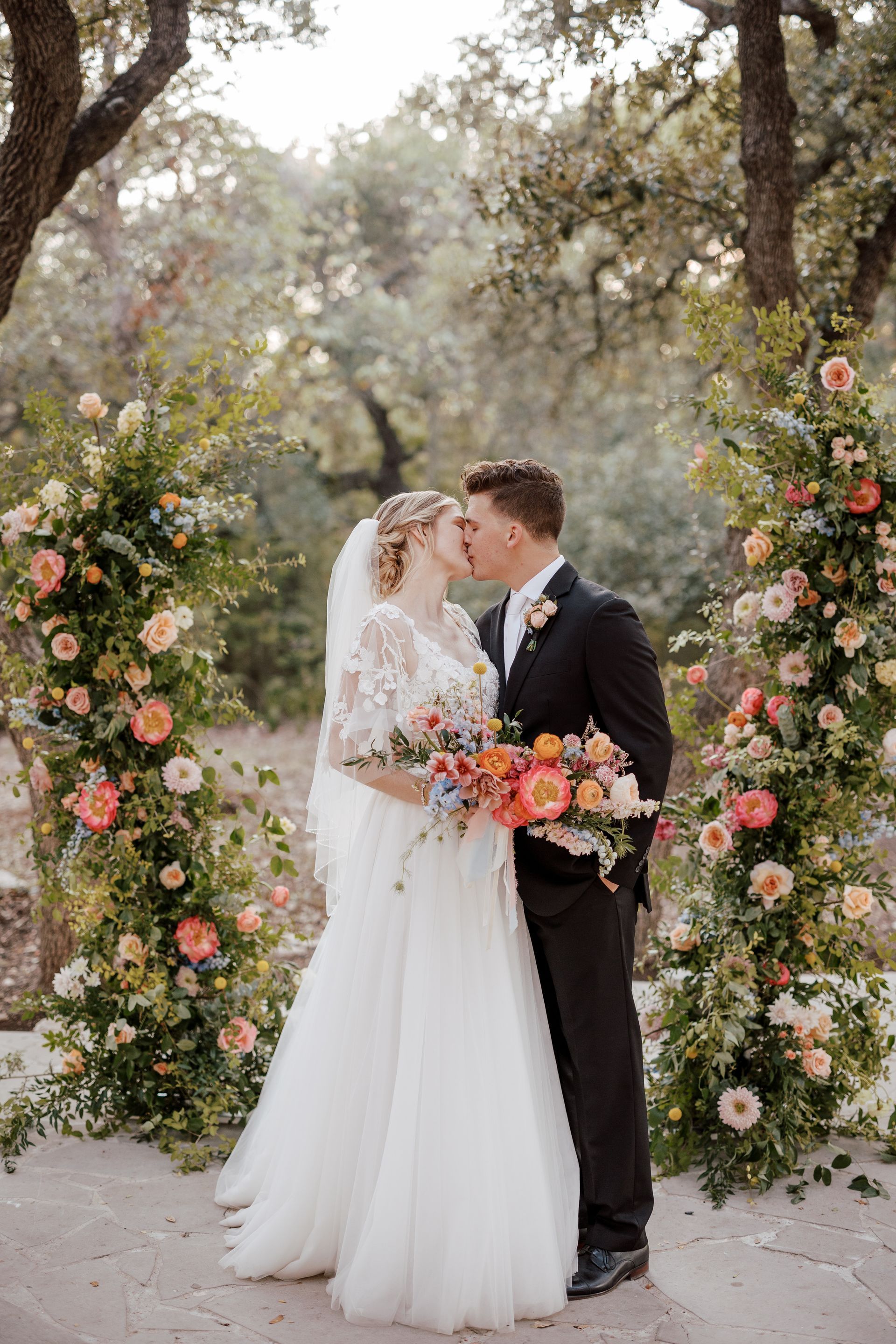 A bride and groom kissing while standing between two large, vibrant floral arrangements in a wooded outdoor setting.