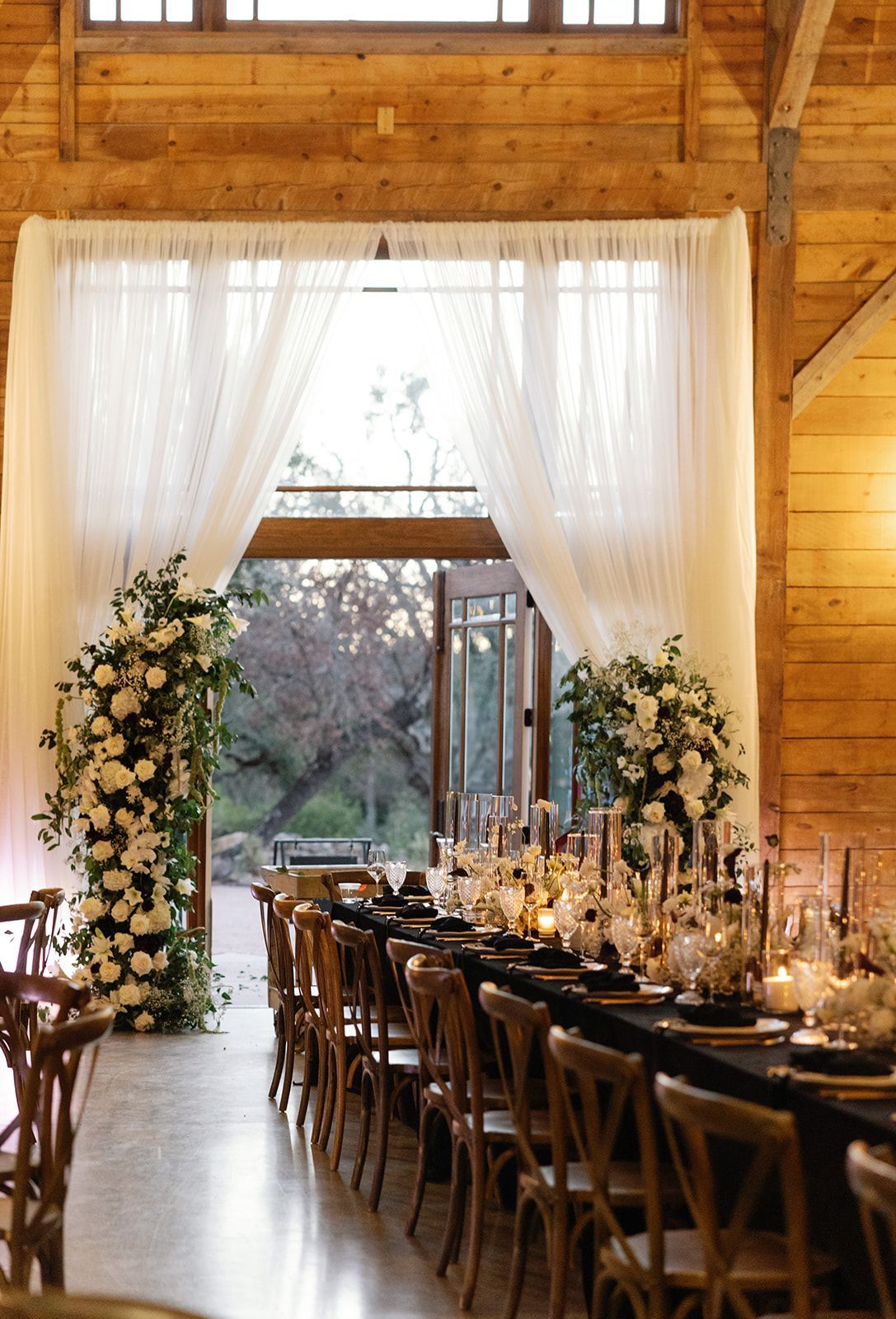 Elegant wedding reception setup in a rustic barn with long table, floral arches, and white drapes