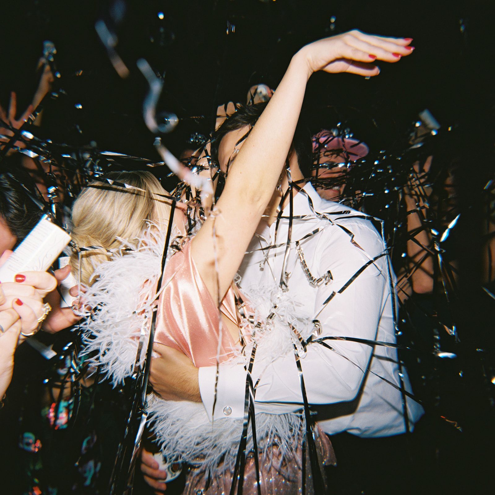 A person wearing a feathered top embraces another individual amidst falling silver confetti in a dark, festive setting.