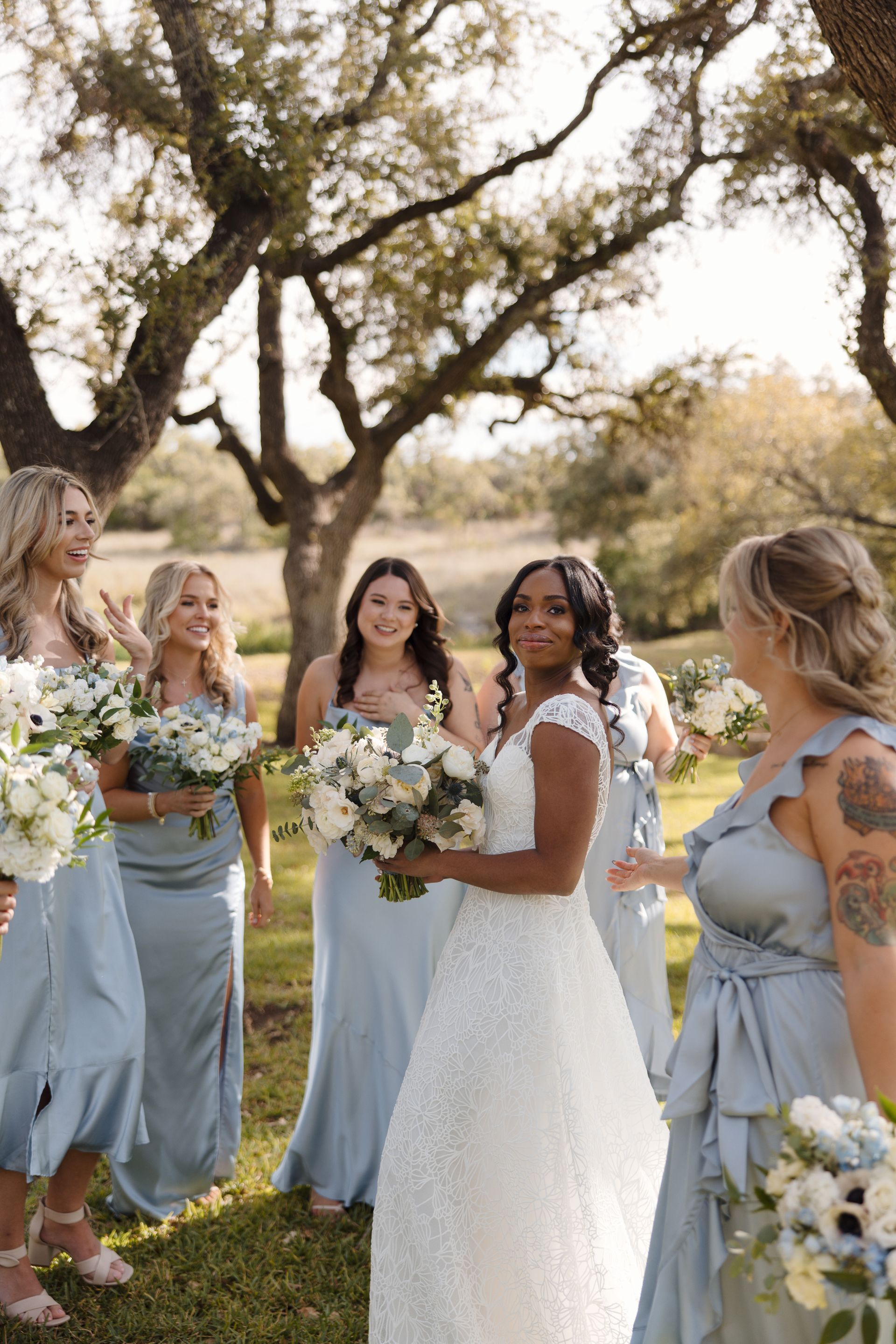 A bride in a white lace gown stands with smiling bridesmaids in light blue dresses, posing outdoors under large trees.