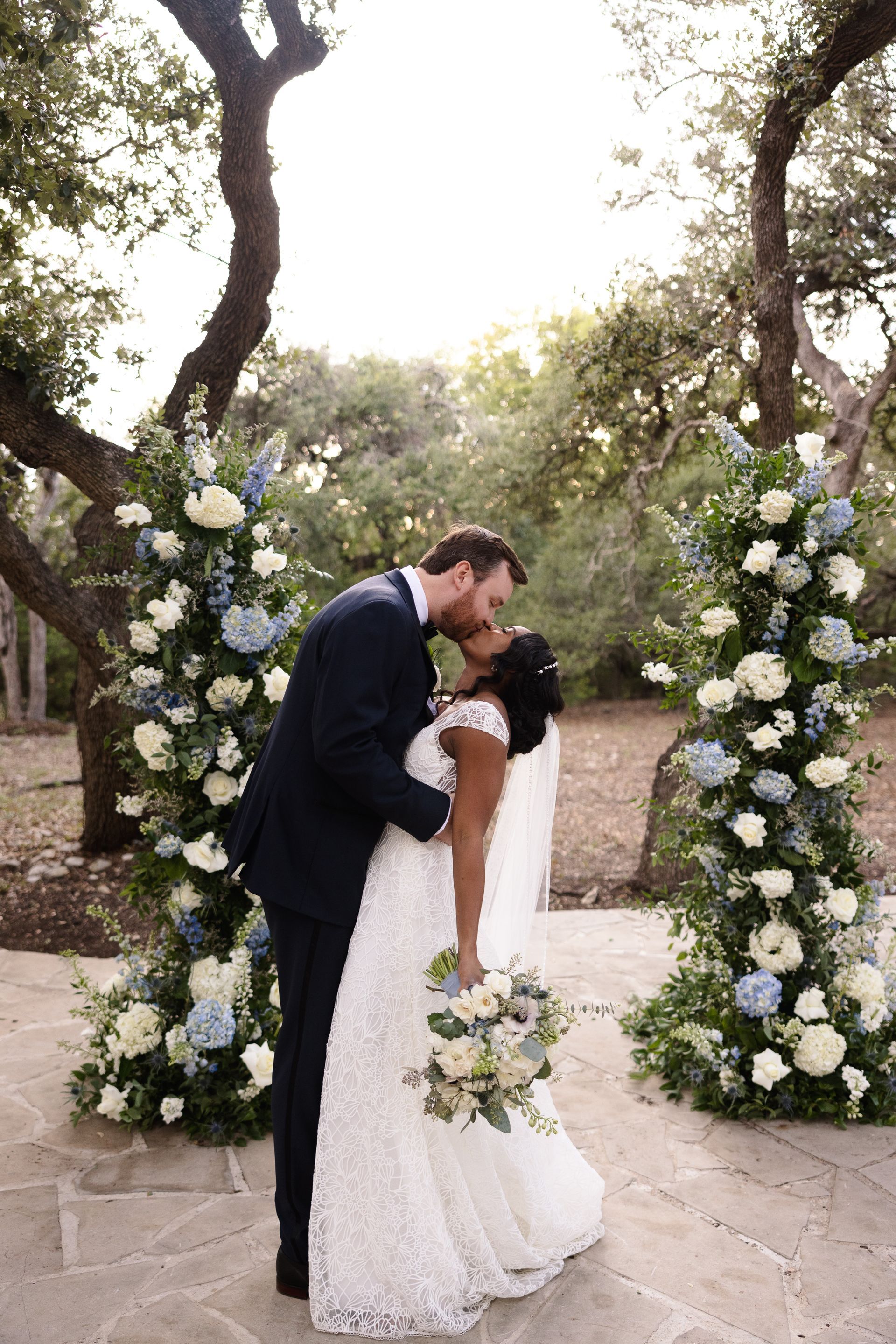 A couple kissing between two tall floral arrangements of white and blue flowers in an outdoor, wooded setting.