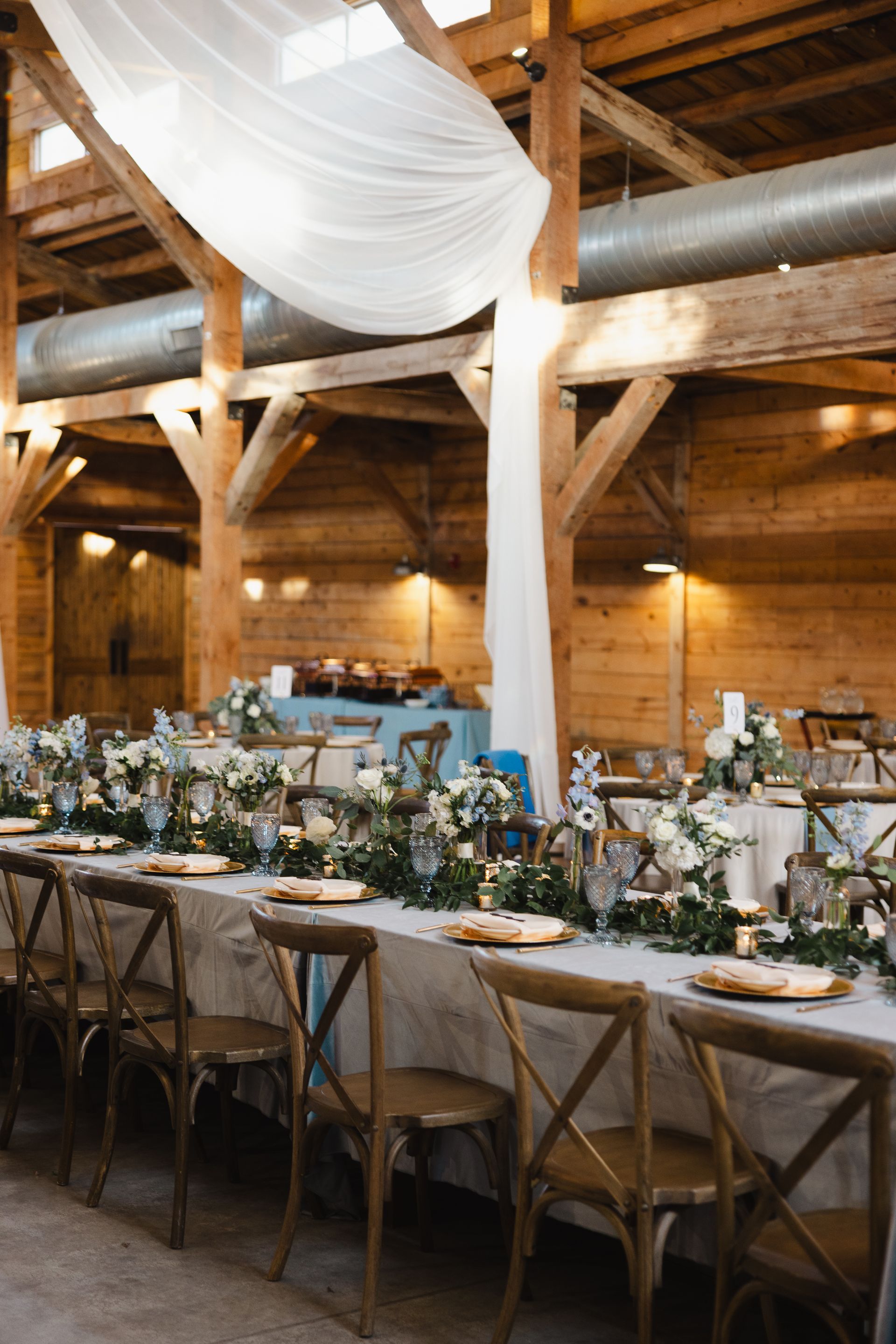 A long table set for a wedding reception in a rustic wooden barn, decorated with white fabric, greenery, and candles.