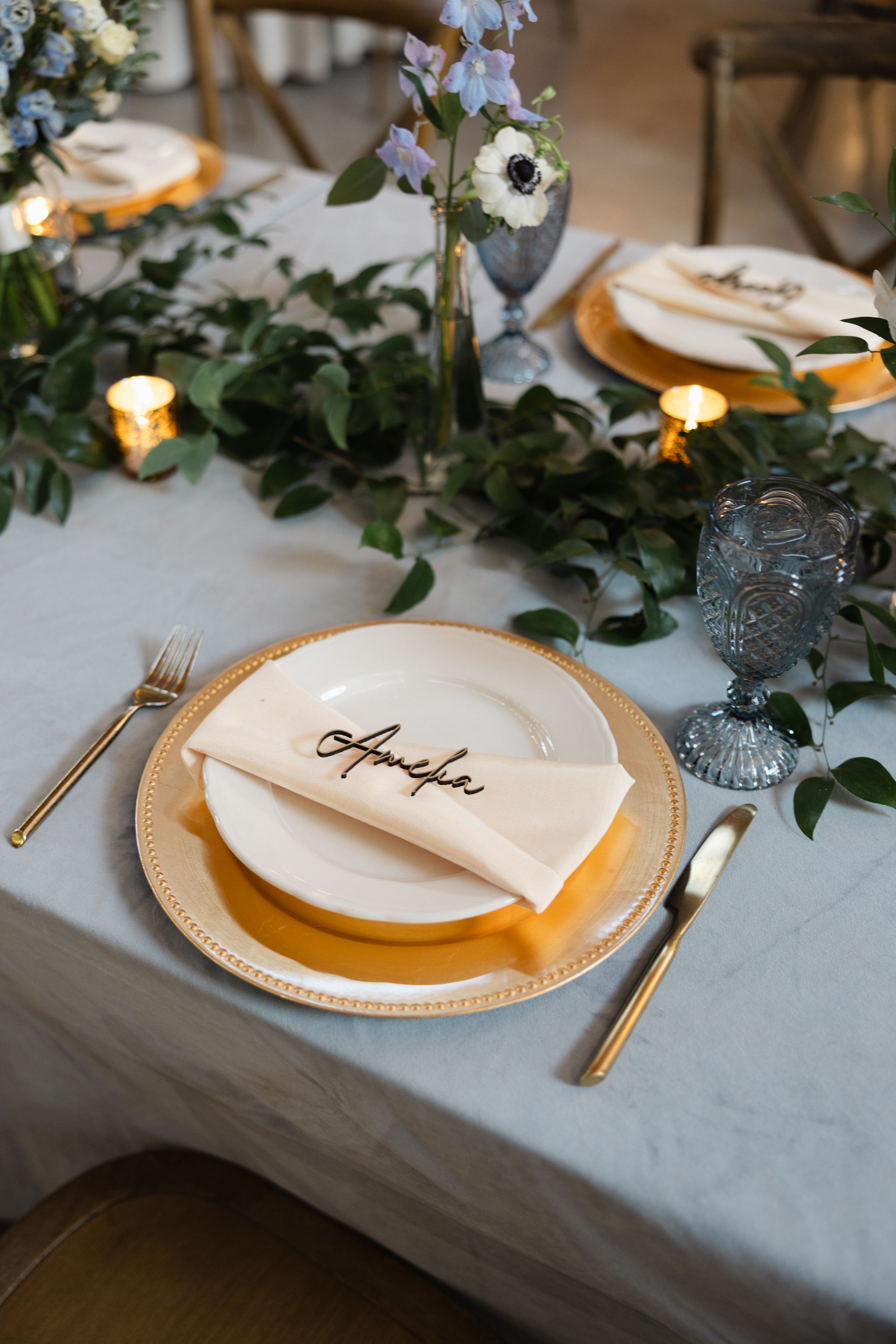 A place setting at a wedding table featuring a white plate, gold charger, floral accents, and a personalized name card.