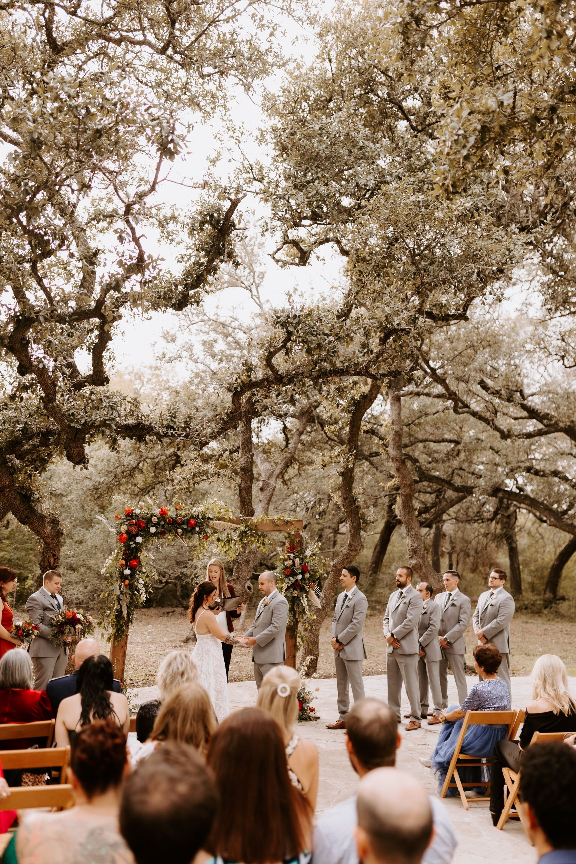 Wedding couple exchanging vows under a floral arch in an outdoor forest setting with guests seated in rows.