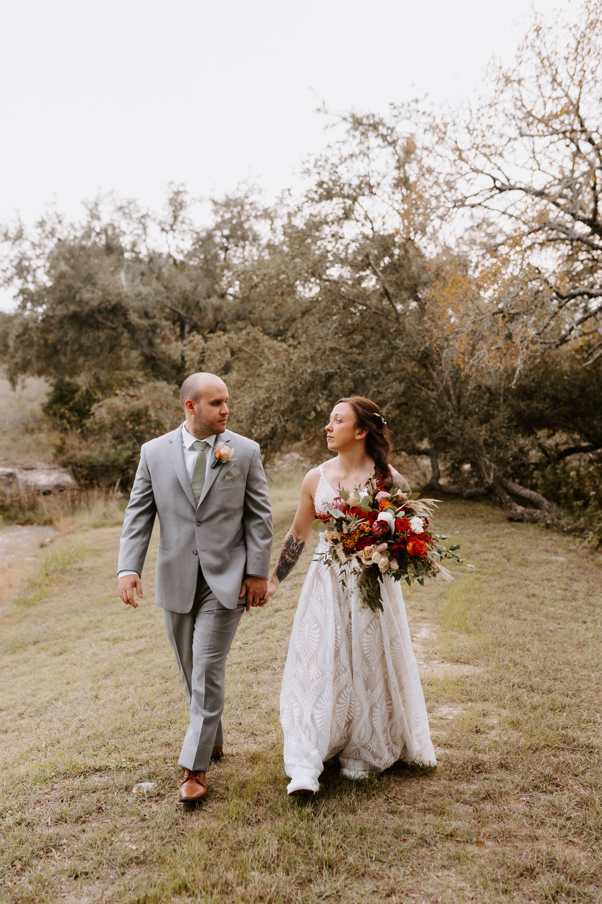 A couple in formal wedding attire walks hand-in-hand through a grassy, wooded area, looking at each other.