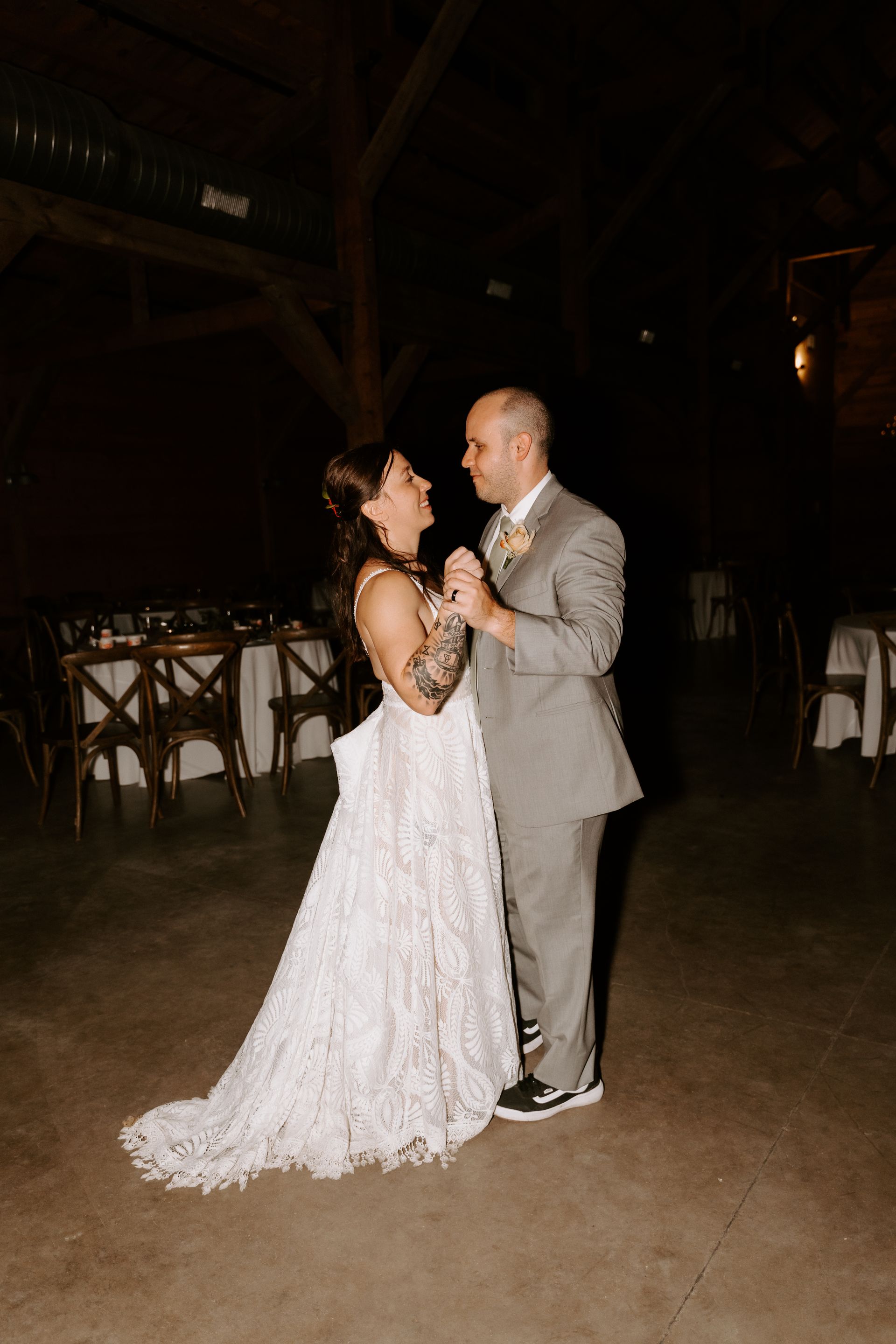 A bride and groom in wedding attire look at each other while slow dancing in a dimly lit, rustic wooden barn interior.