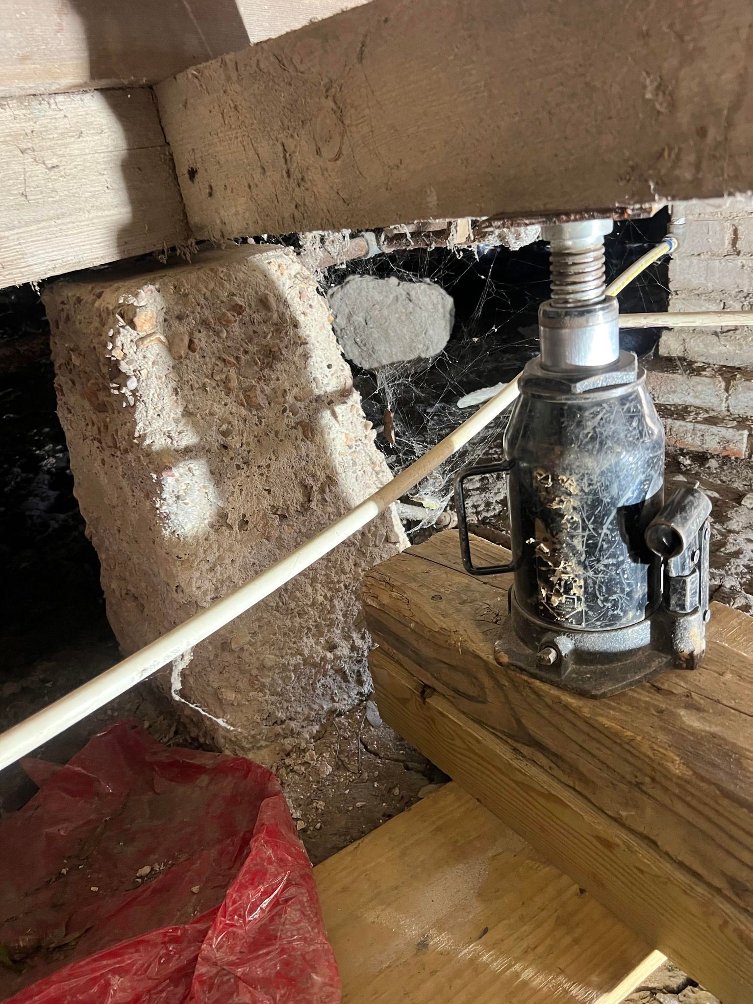 Jack supporting a wooden beam in a cramped crawlspace with brick and stone walls.