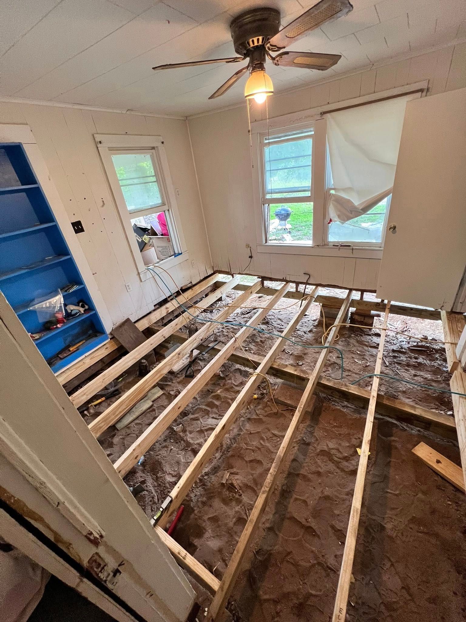 Room under renovation with exposed floor joists, debris, and a ceiling fan in a small bedroom