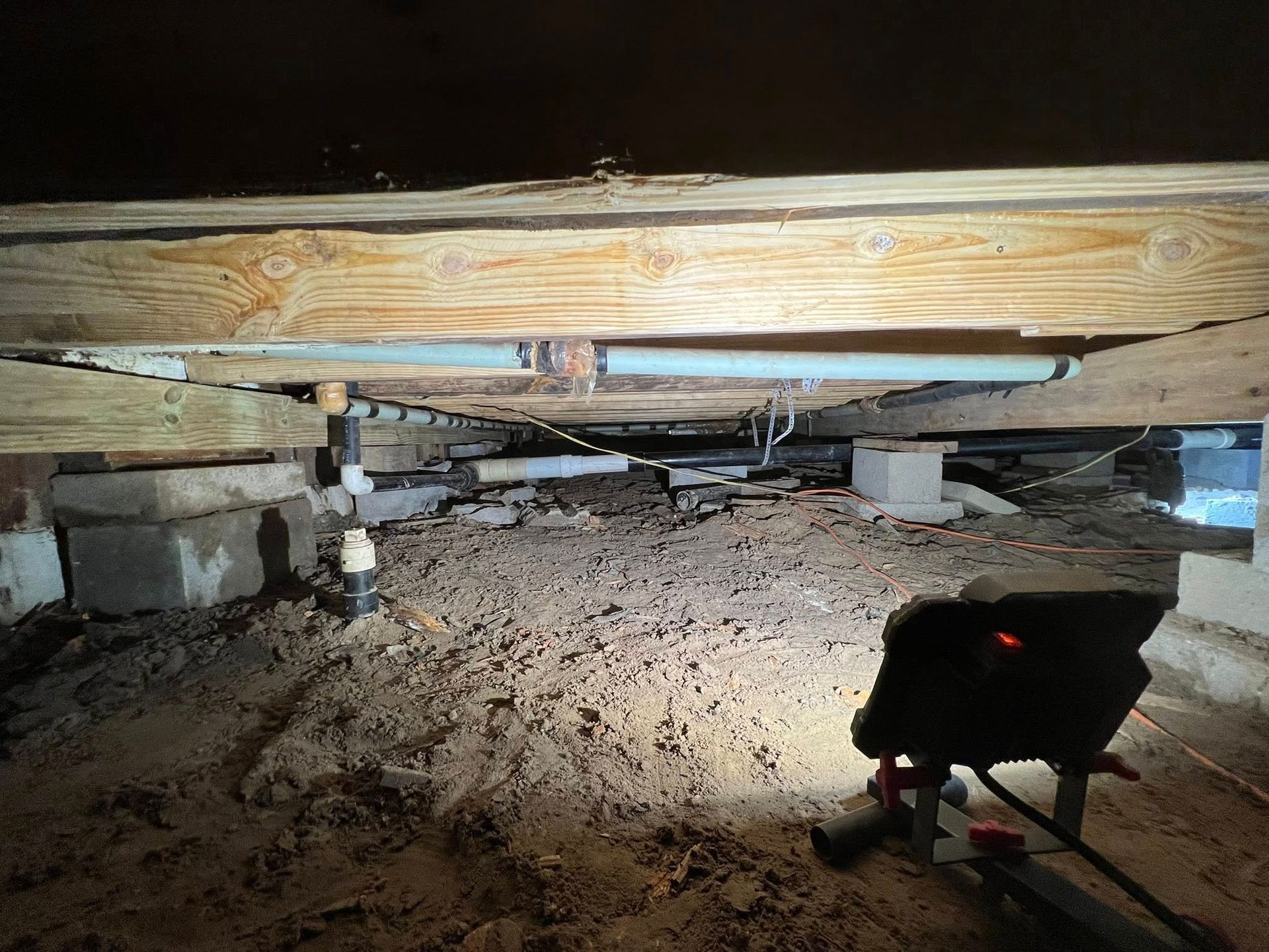 Crawling under a low wooden floor with dirt and pipes visible, lit by a small flashlight.