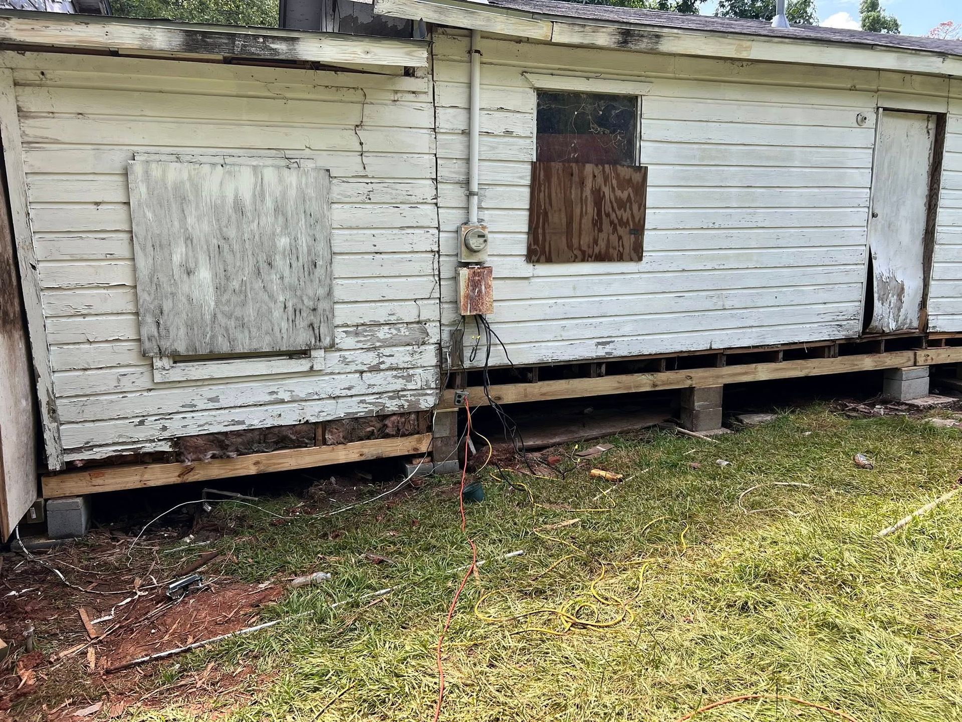 Dilapidated white wooden shack with boarded-up windows, raised on blocks over a grassy yard