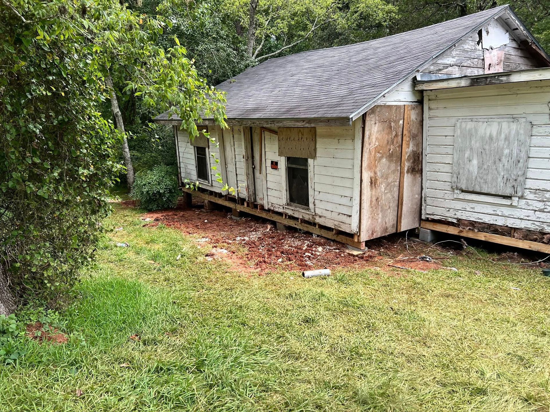 Old, weathered white house with boarded windows and overgrown yard under trees