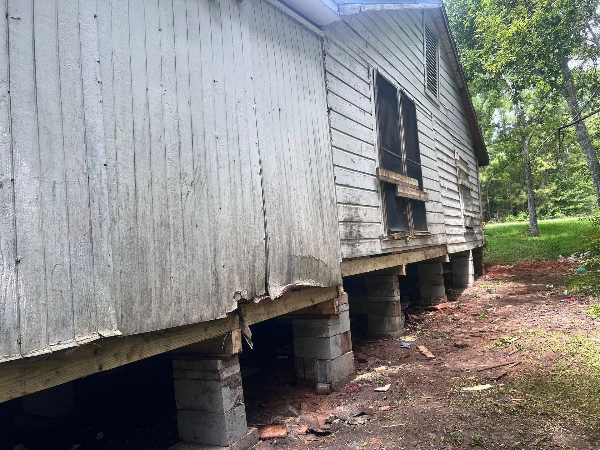 Old wooden house on blocks with a sagging foundation and damaged siding beside a wooded yard.