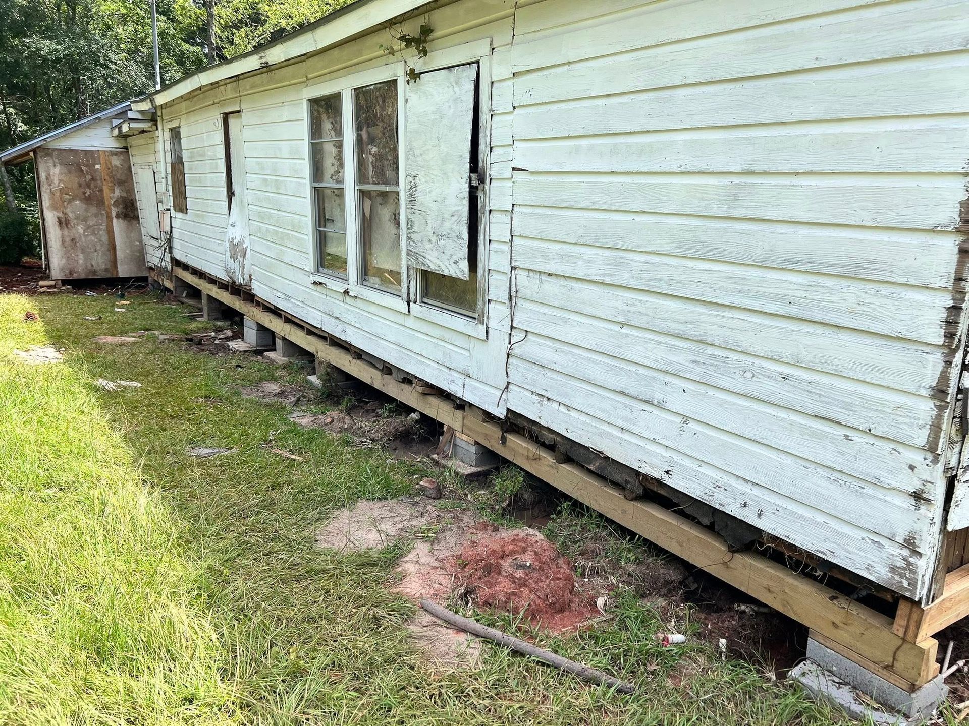 White house exterior with damaged siding, exposed support beams, and broken windows beneath a sloping roof