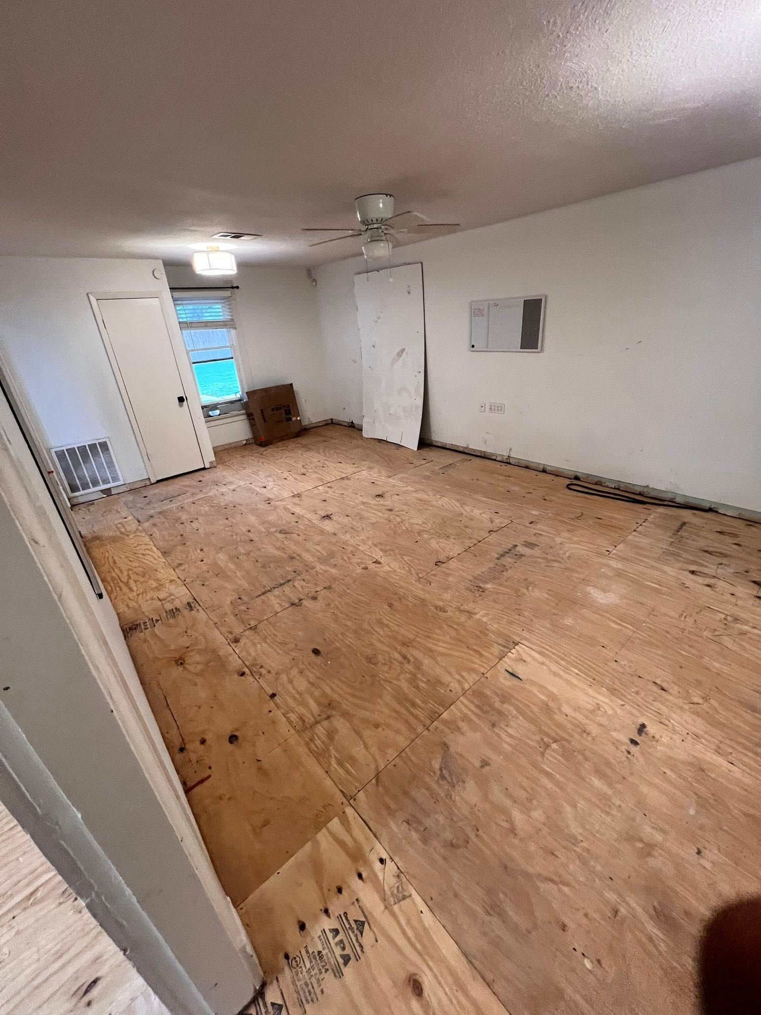 Empty unfurnished room with plywood floor, white walls, ceiling fan, and open doorway to a lit adjoining space