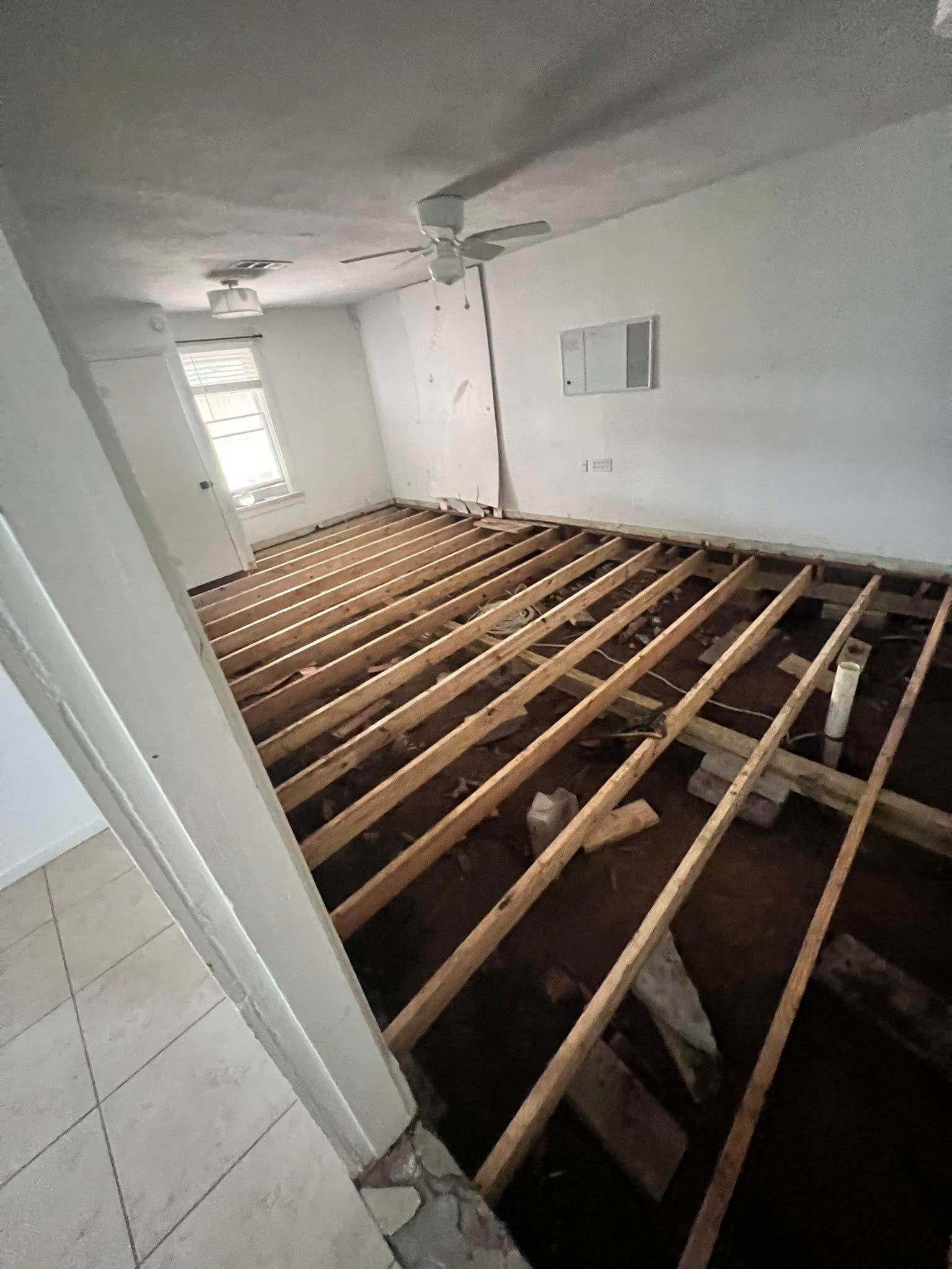 Interior room with exposed floor joists, white walls, and a ceiling fan near a window.